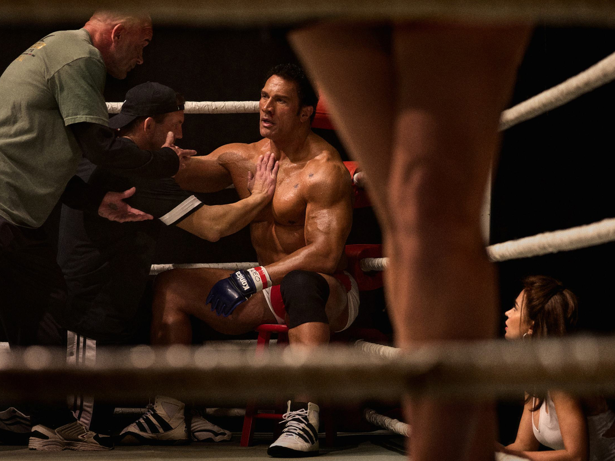 Shirtless boxer sitting in ring corner receiving medical attention from trainer whilst another person tends to him between rounds.
