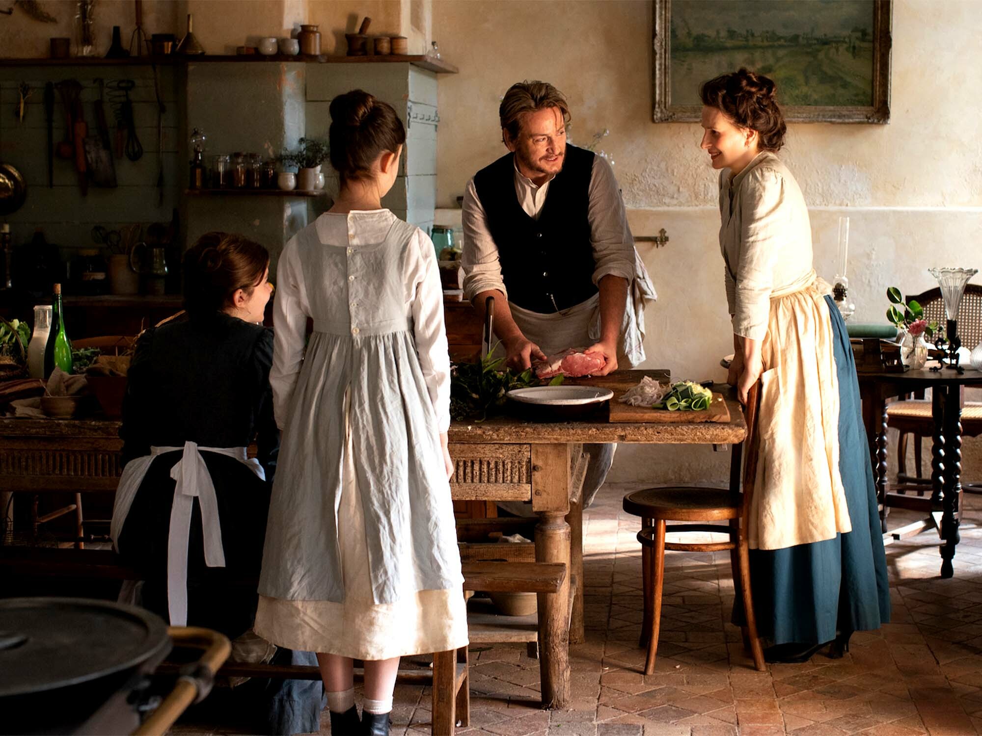 Three women in period costumes stand in a rustic kitchen, preparing food on a wooden table.