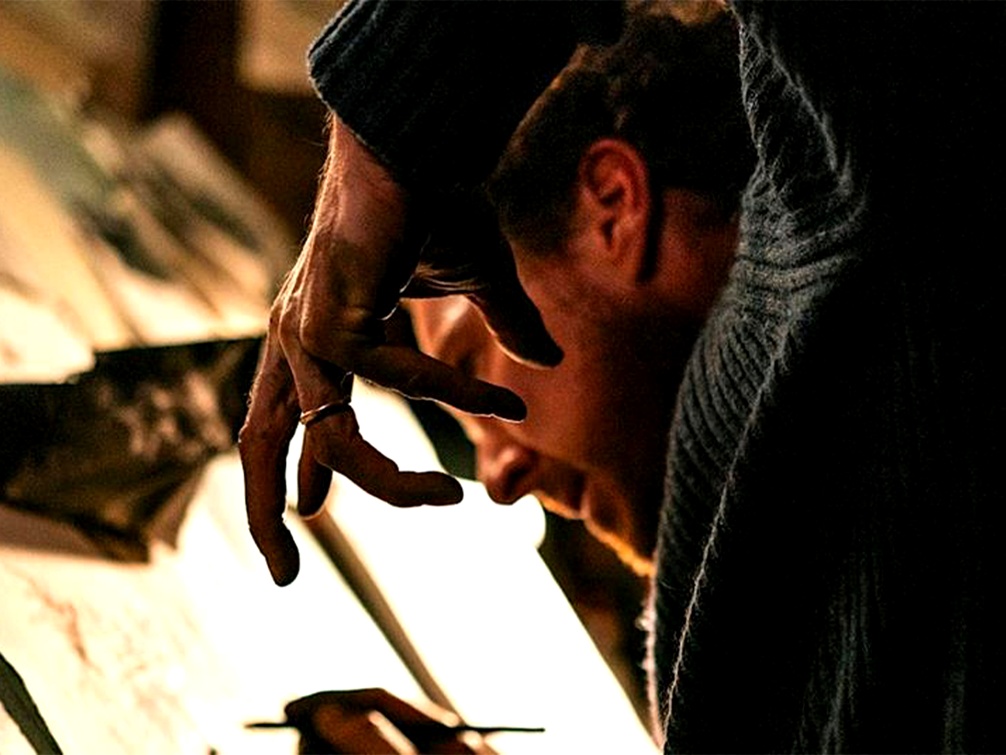 Man in grey jumper working with hands on wooden surface, warm lighting illuminating his focused profile against blurred background.