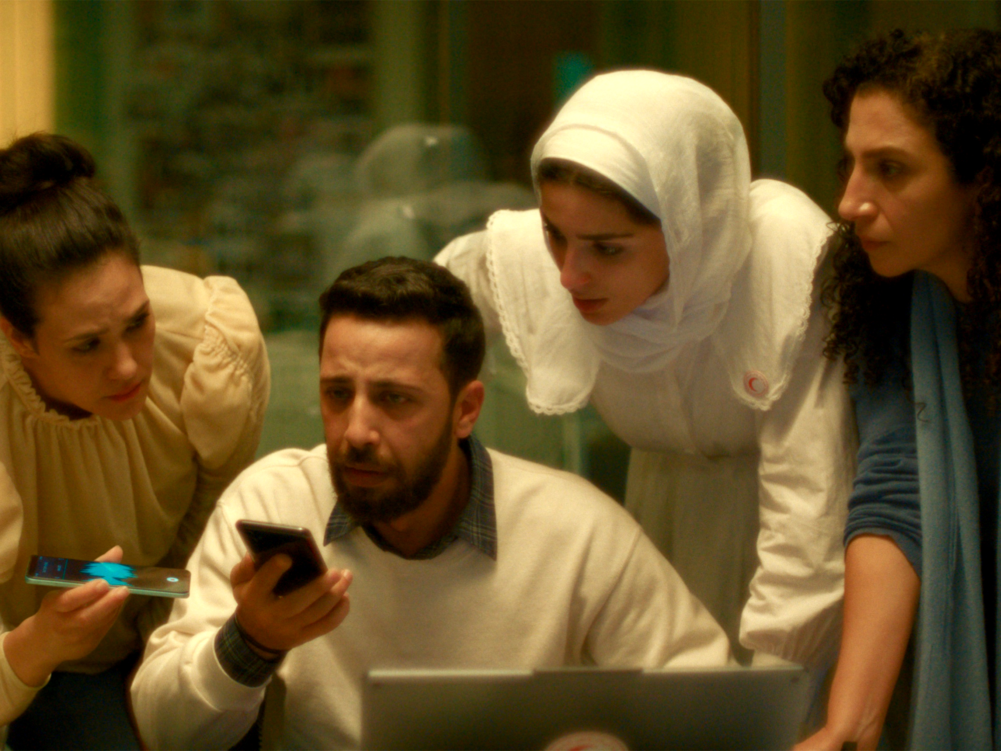 Five people gathered around a laptop computer, with a bearded man in white shirt at centre holding a mobile phone.