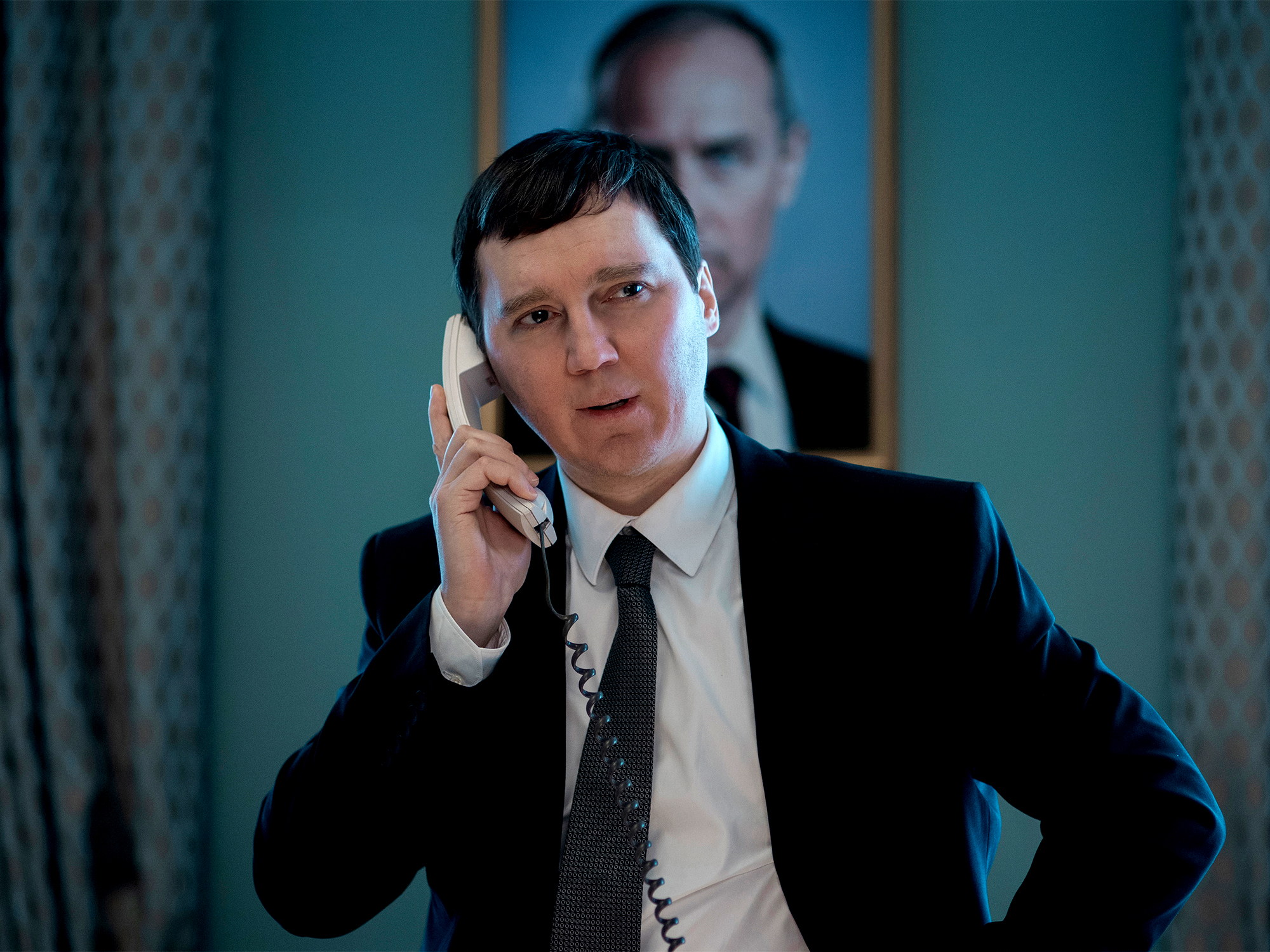 Man in dark suit speaking on phone in formal interior setting, with blurred figure in background.