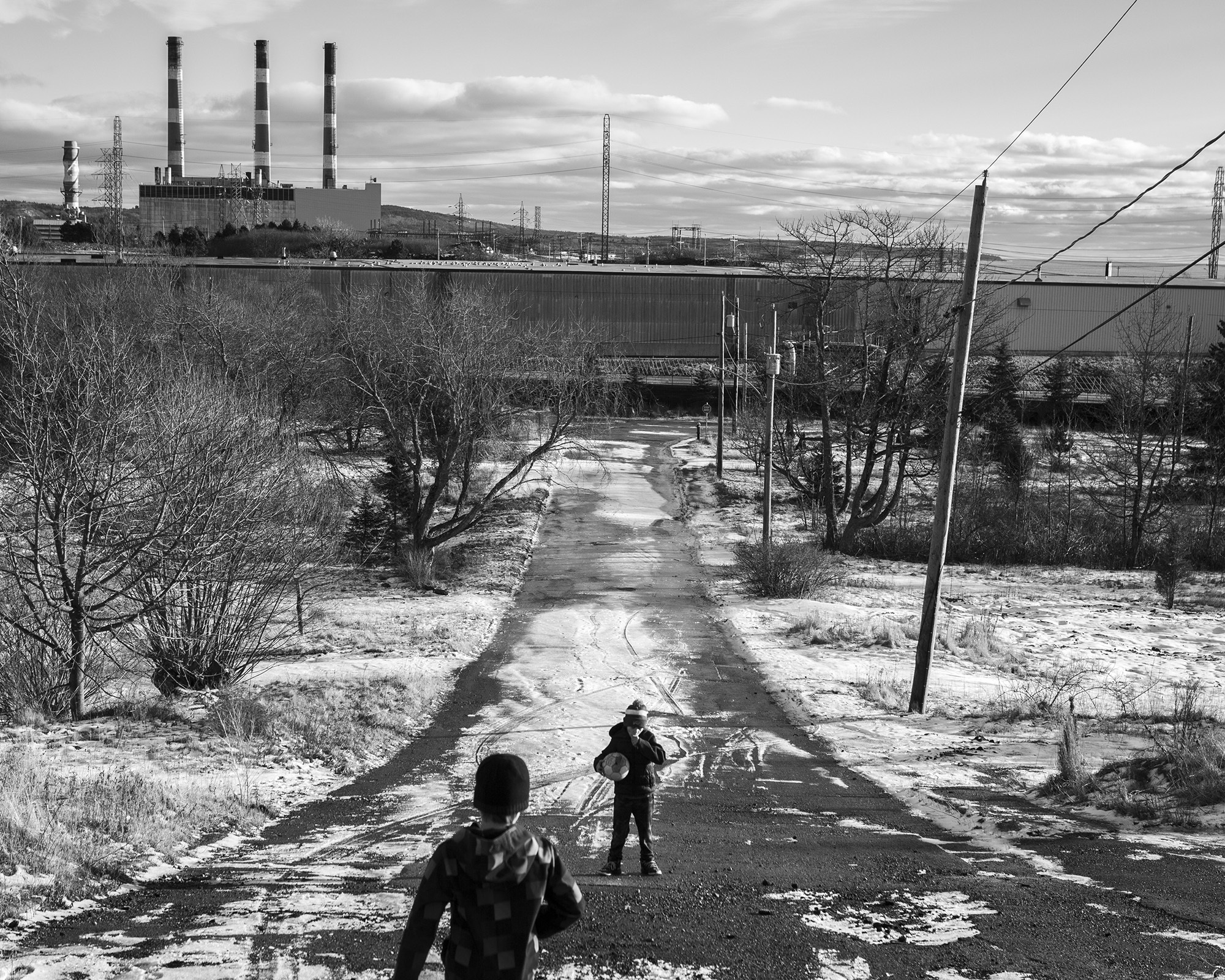 Snowy path leading to industrial plant with tall chimneys, overcast sky, bare trees.