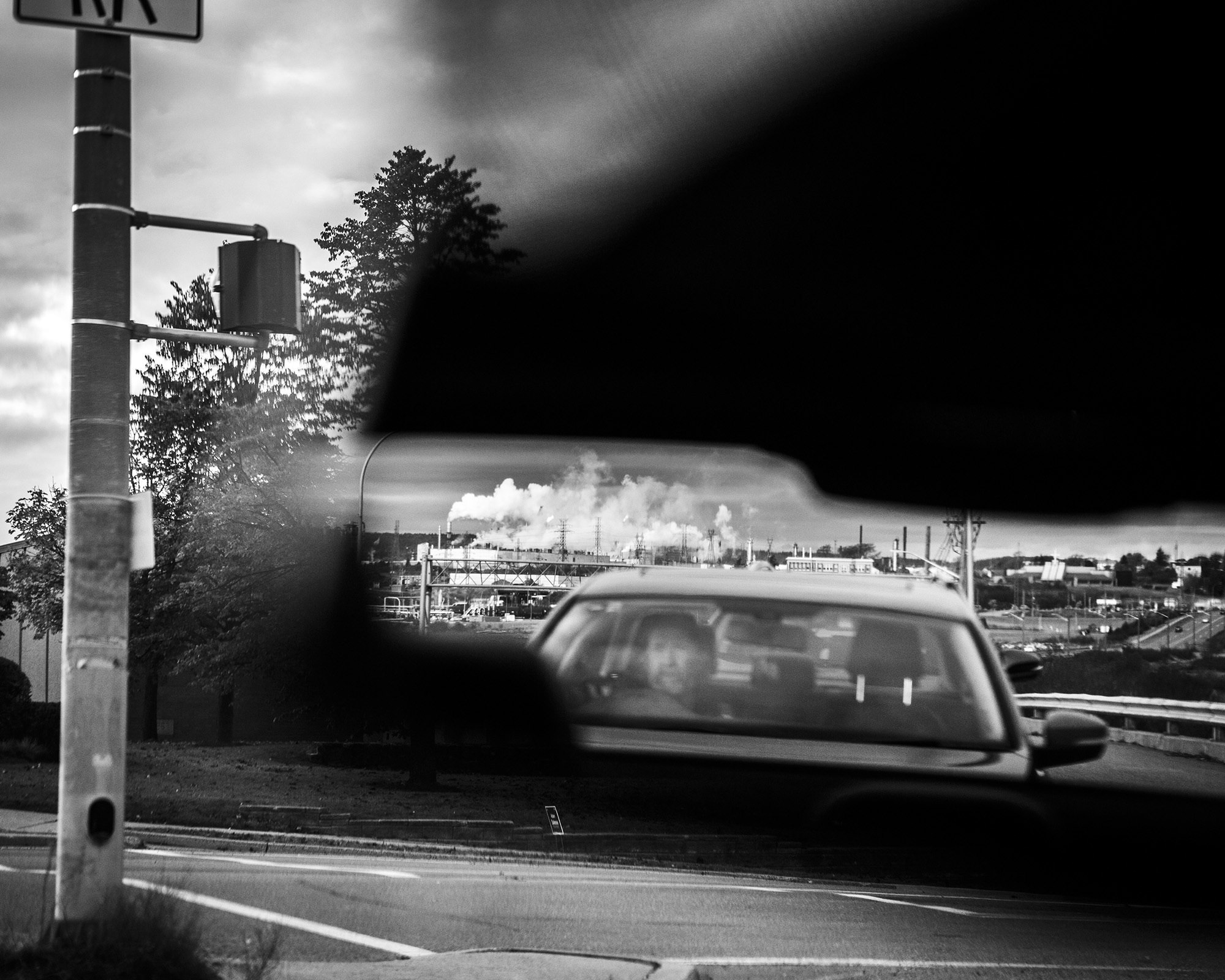 Black and white image of a city skyline in the distance, with a blurred car in the foreground and a traffic light pole on the left.