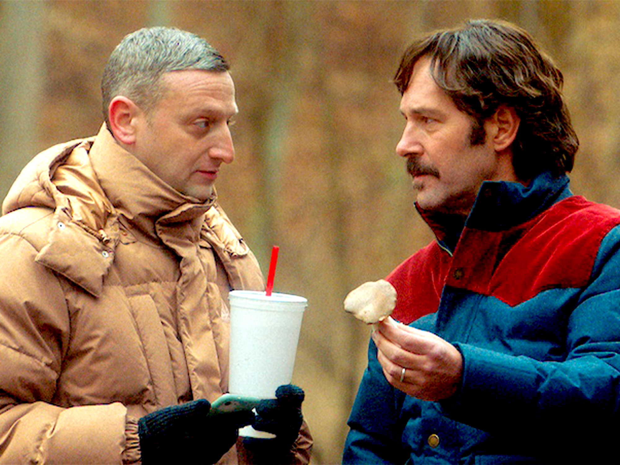 Two men in conversation outdoors, one in beige puffer jacket holding white cup with red straw, other in red and blue jacket eating.
