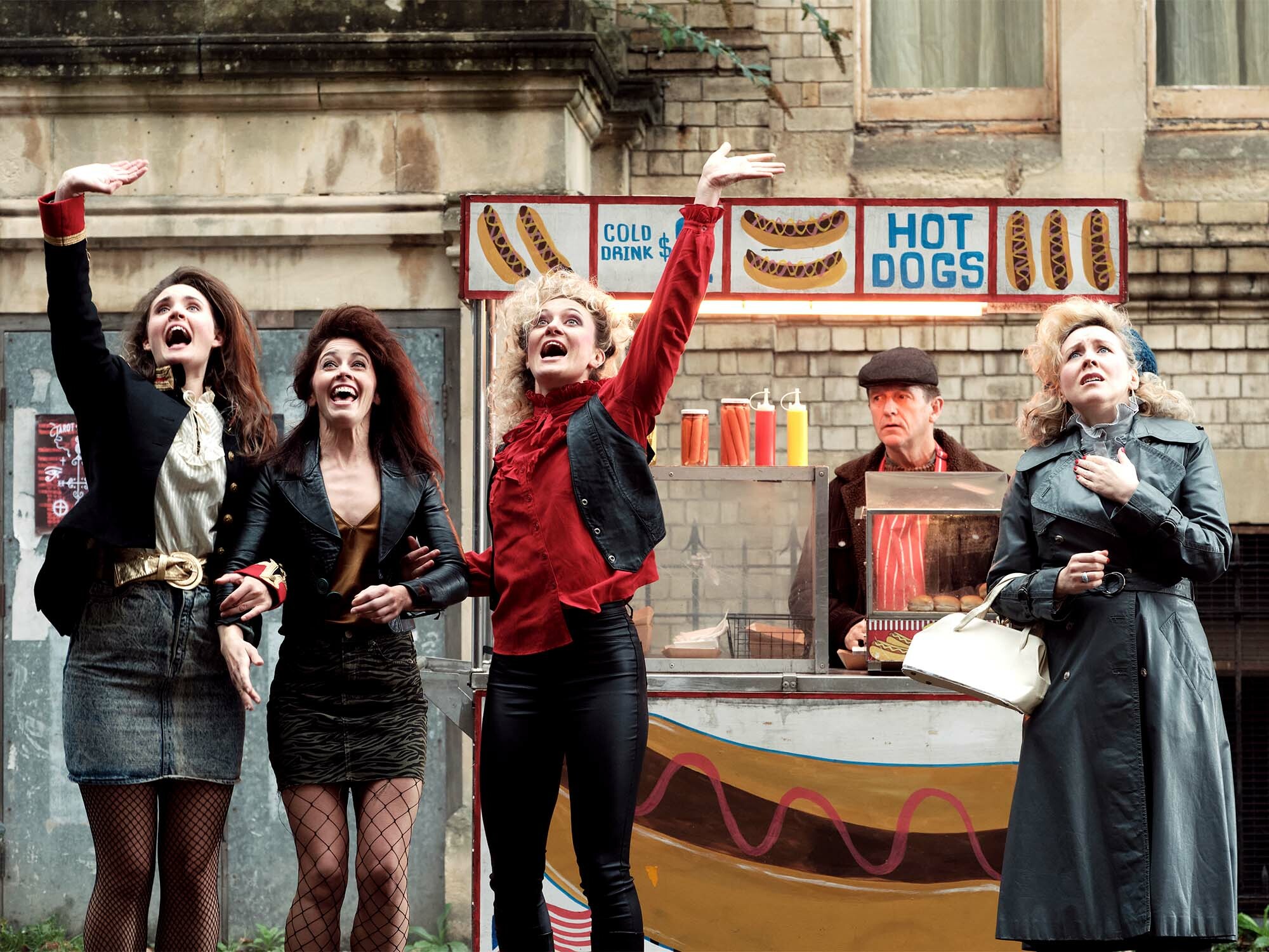 Five young women posing outside a hot dog stand, some with arms raised. The women wear dark clothing, jackets, and one has a red jacket. In the background, a sign advertises "Hot Dogs".