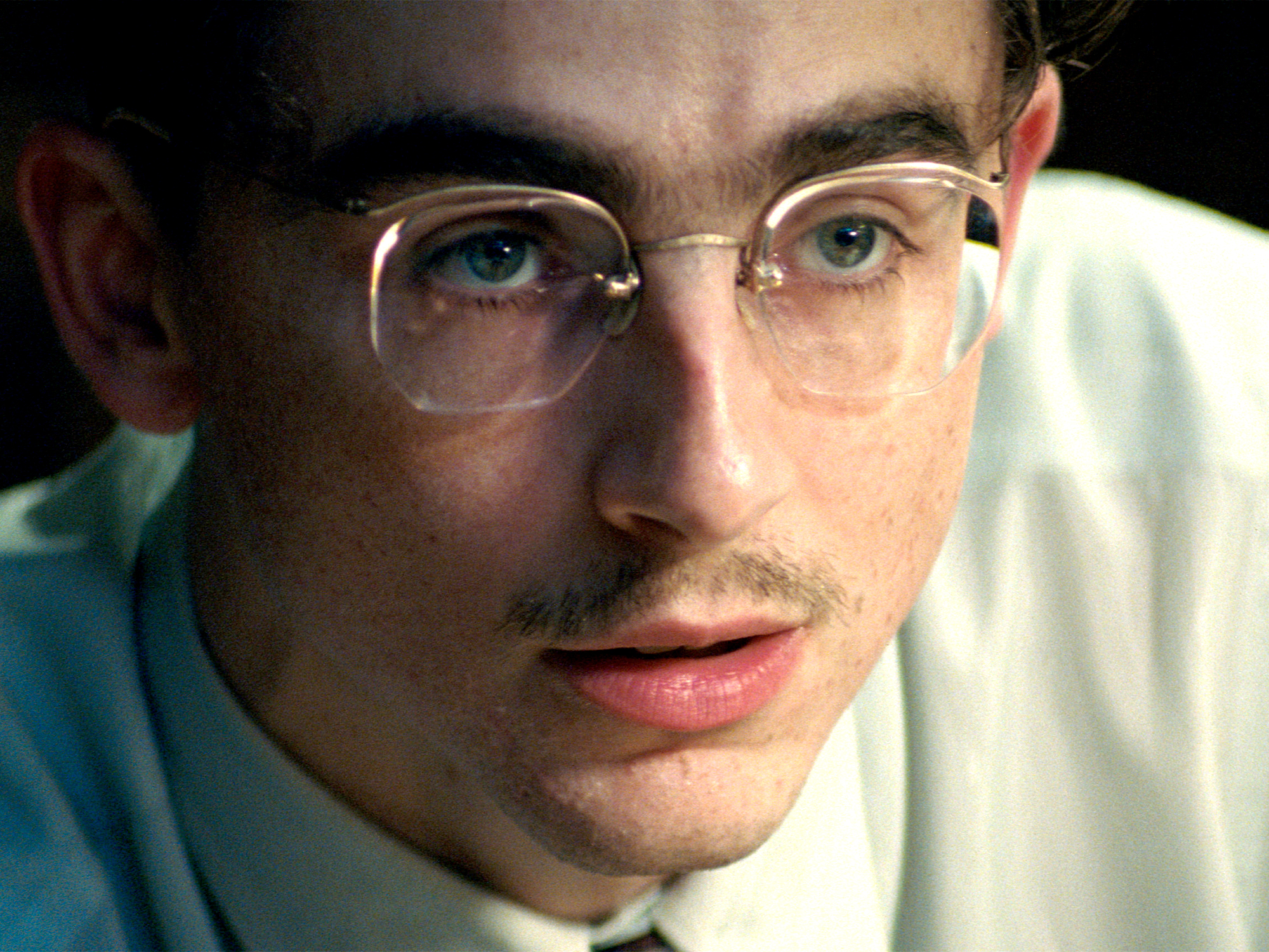 Close-up of a man with wire-rimmed glasses, blue eyes, and facial hair wearing a white shirt against a dark background.