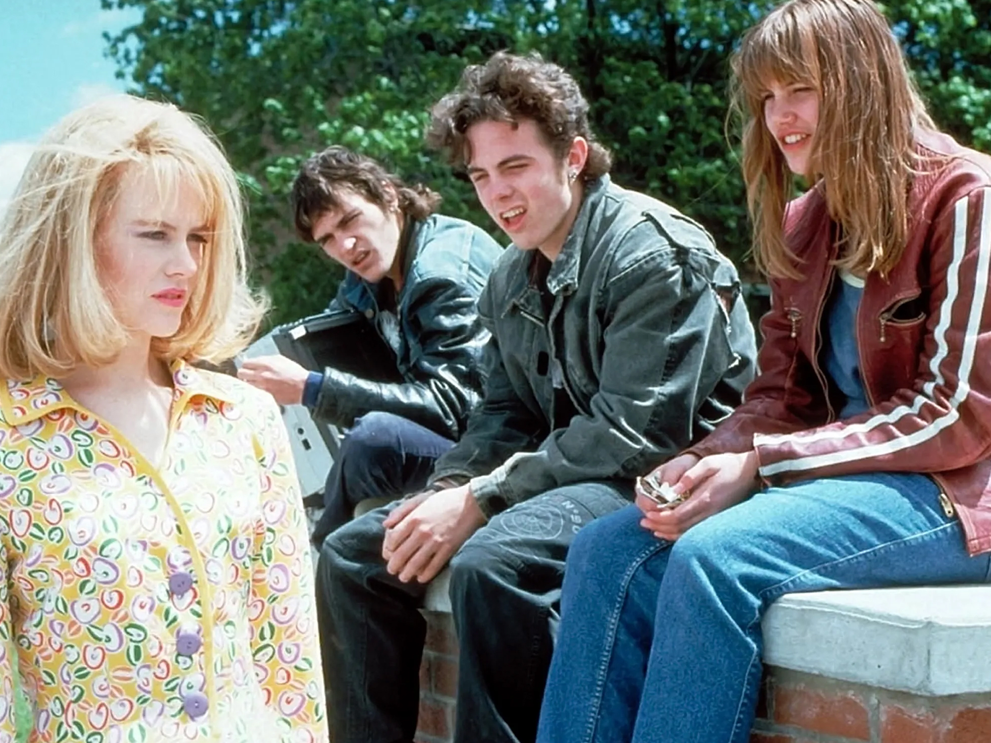 Nicole Kidman dressed in a yellow patterned blouse stands in front of a wall. Sitting on the wall are a young Joaquin Phoenix, Casey Affleck and Busy Phillips, who are all dressed in jeans and jackets.
