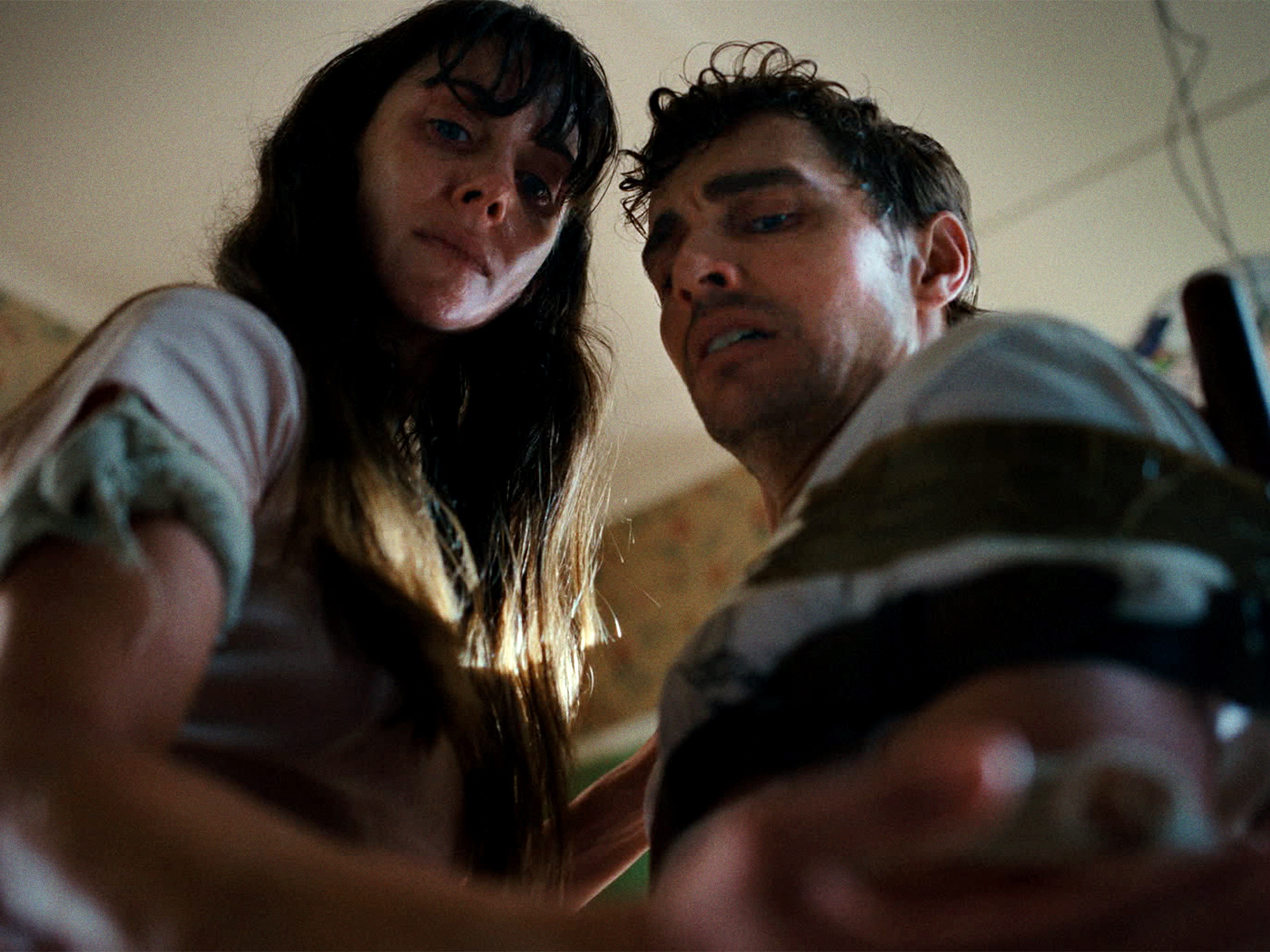 Woman with long dark hair and man with curly hair looking down at camera in indoor setting.