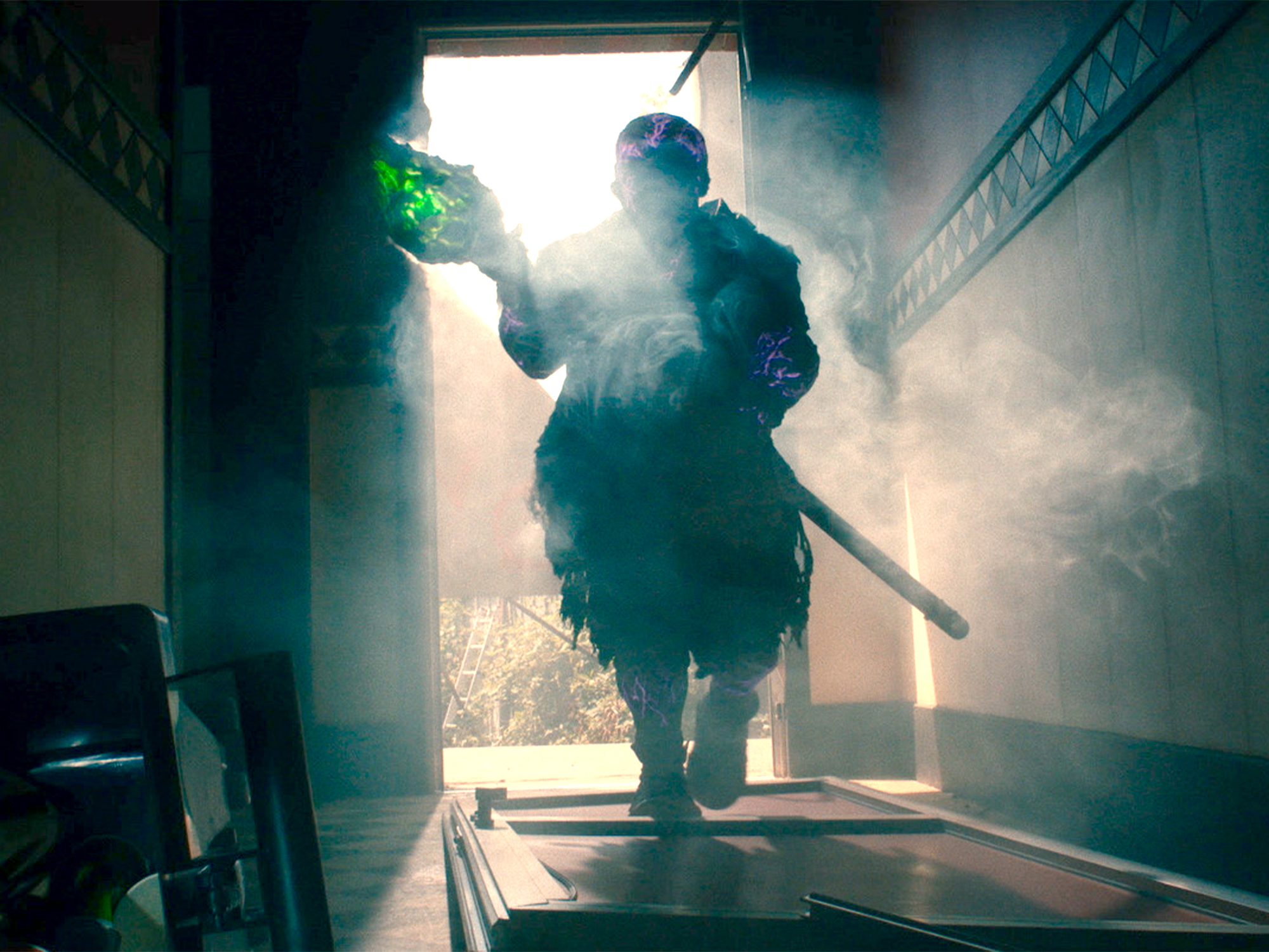 Firefighter in protective gear walks through smoky doorway with bright sunlight streaming from behind, creating dramatic silhouette effect.