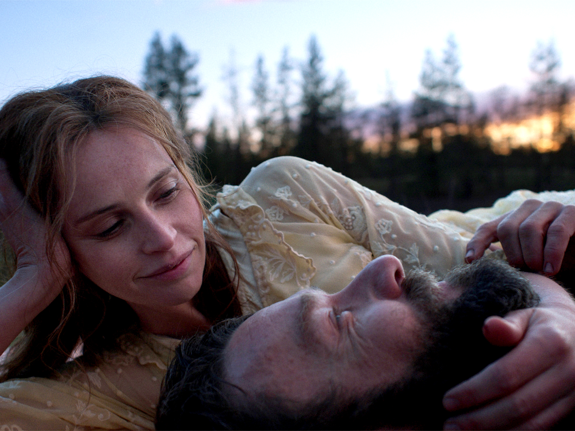 Woman with blonde hair looking down at man with dark hair lying on ground at dusk, trees and warm light in background.