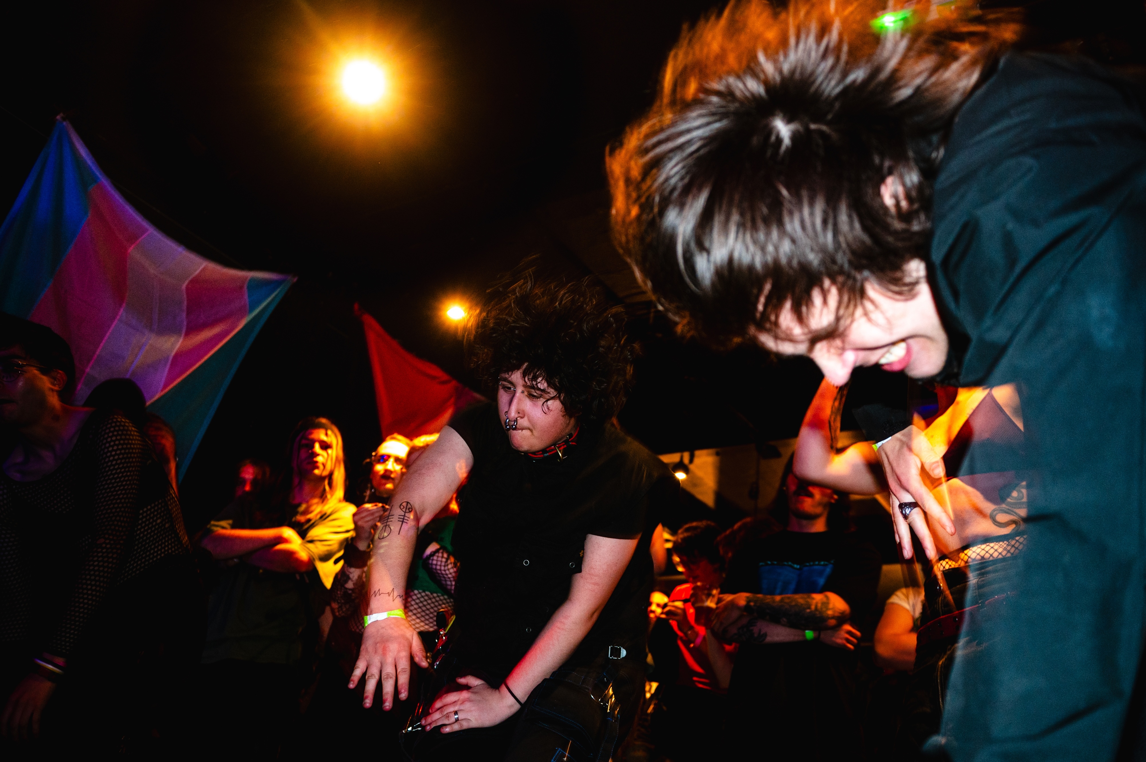 Person with spiky black hair leaning over DJ equipment at night venue with orange lighting and colourful banners in background.