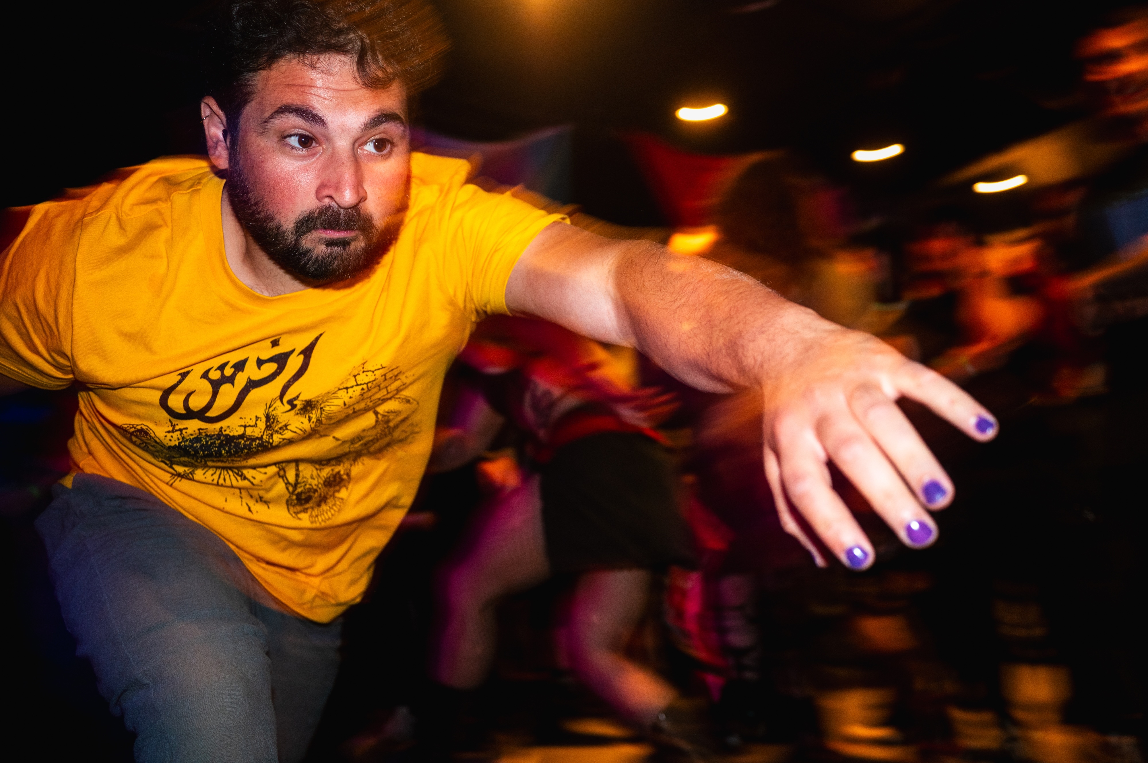 Bearded man in yellow t-shirt with arms outstretched, motion blur in background with warm orange and red lighting.