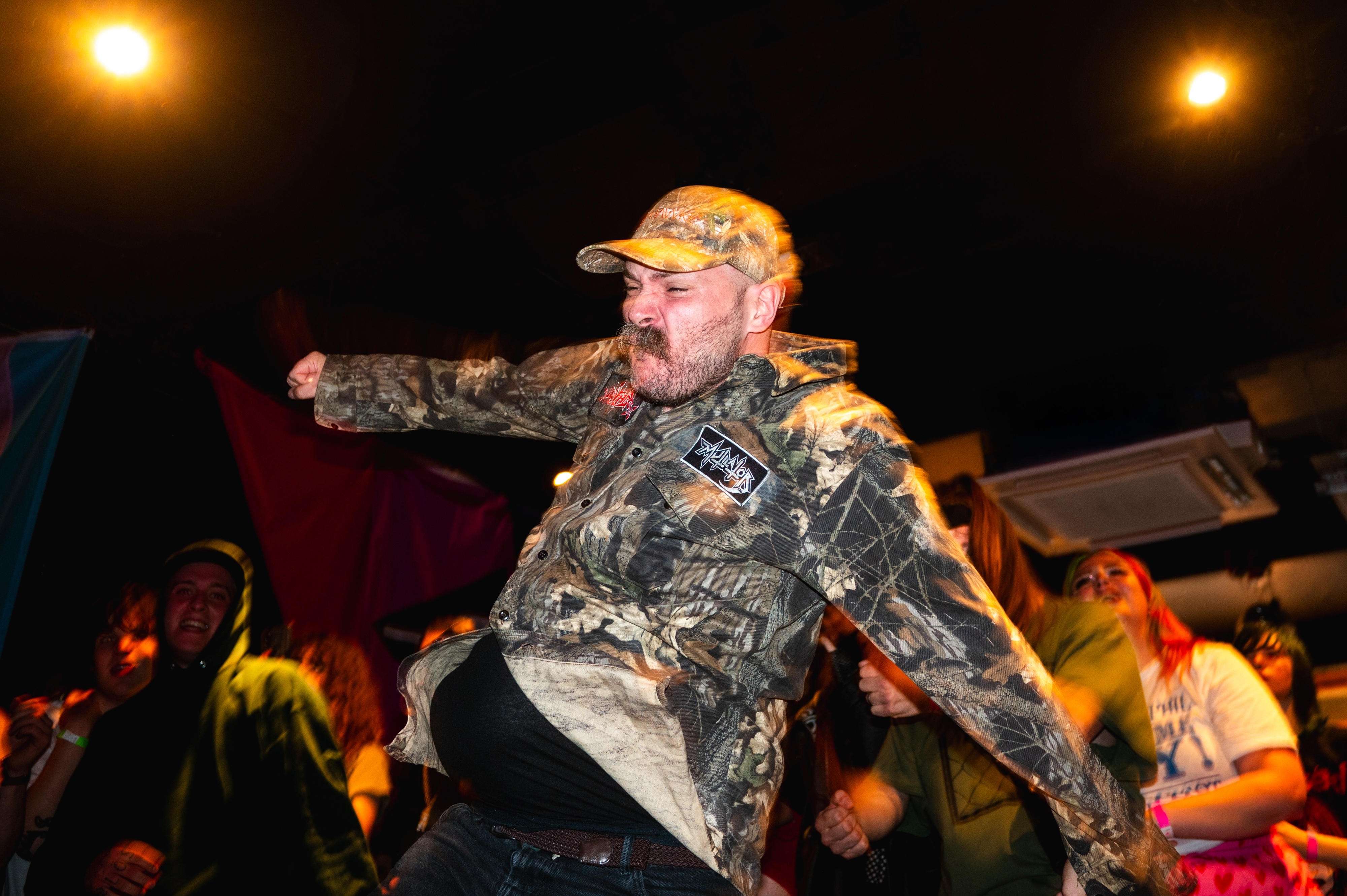 Man in camouflage jacket and cap performing on stage with orange stage lights and crowd in background.