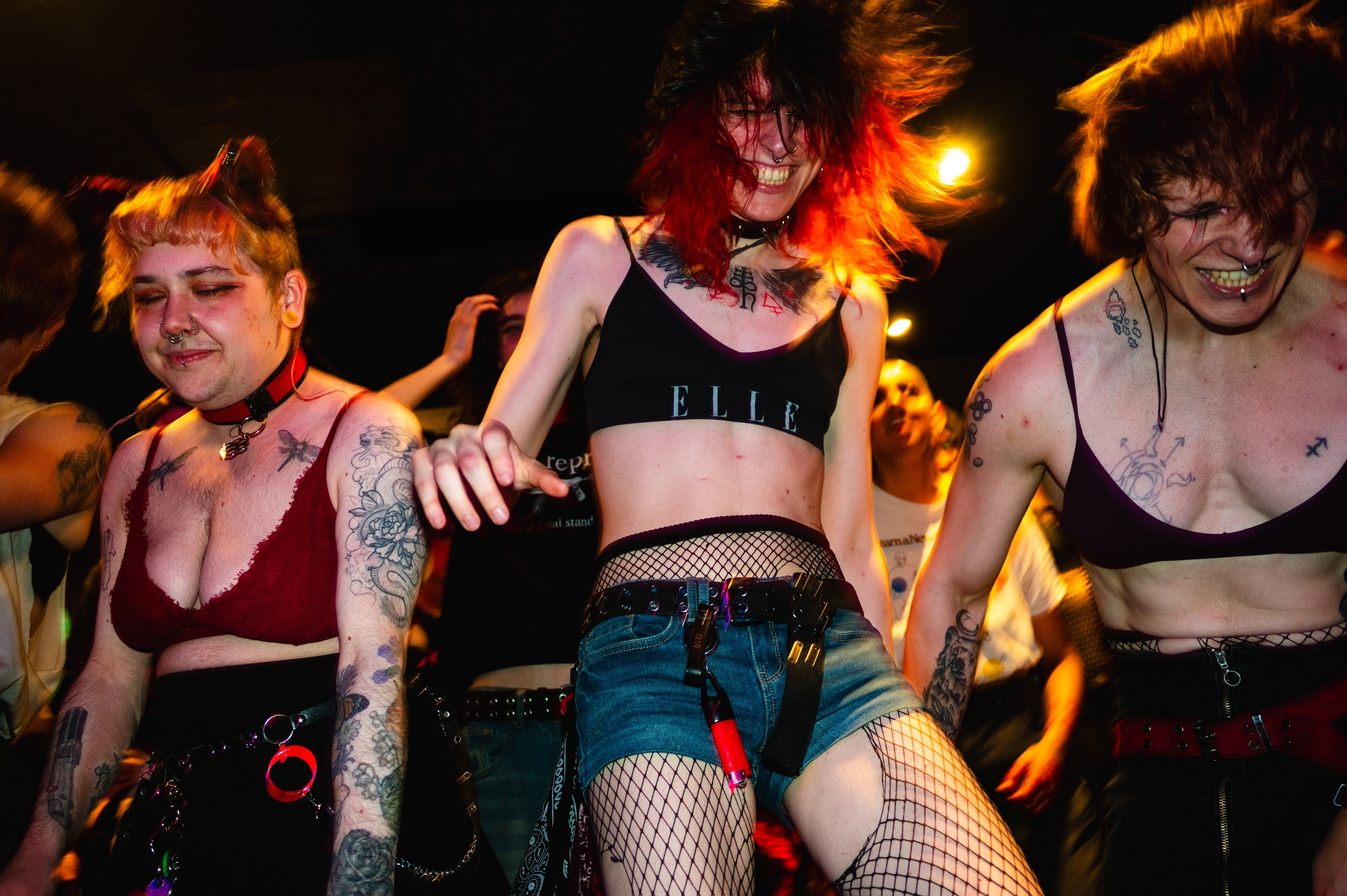 Three performers in alternative fashion with fishnet stockings, crop tops, and denim shorts dancing under warm orange stage lighting.