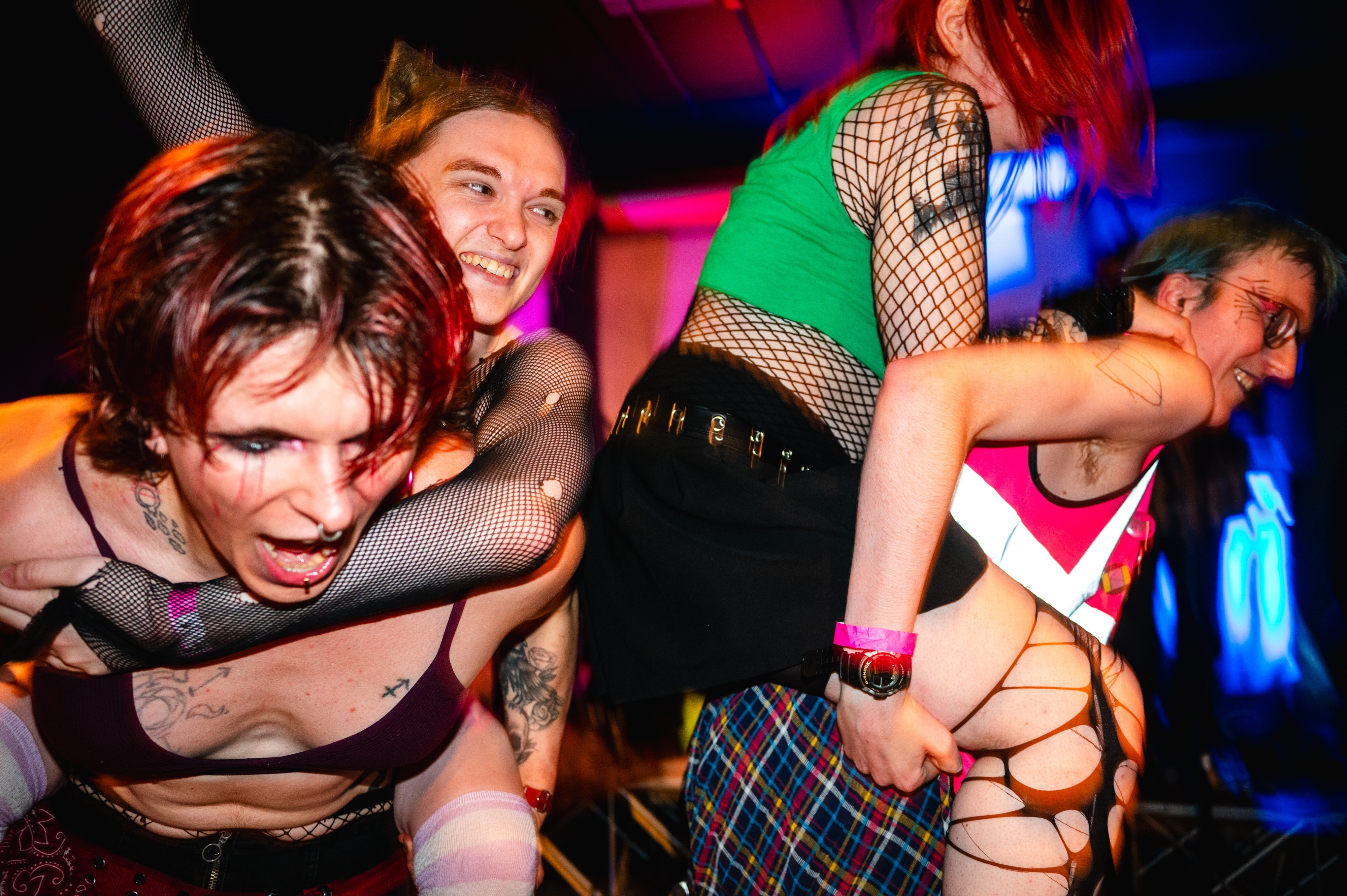 People dancing at nightclub with colourful lighting, wearing fishnet stockings, plaid skirts, and alternative fashion styles.