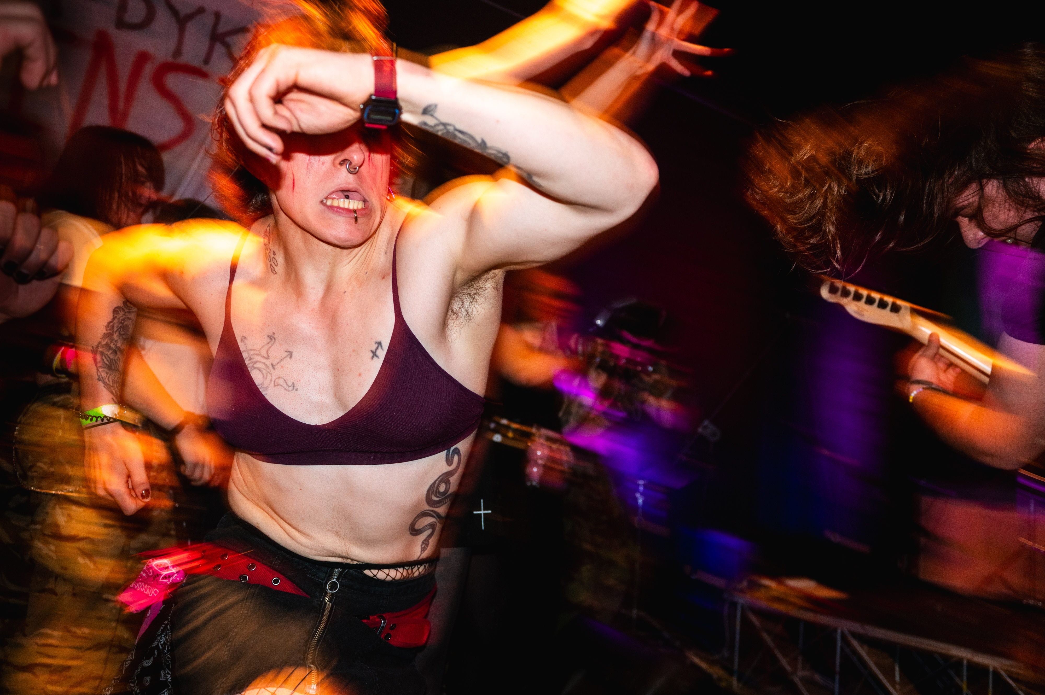 Woman in black bra and dark trousers dancing with arm raised, surrounded by blurred figures in orange and purple lighting.