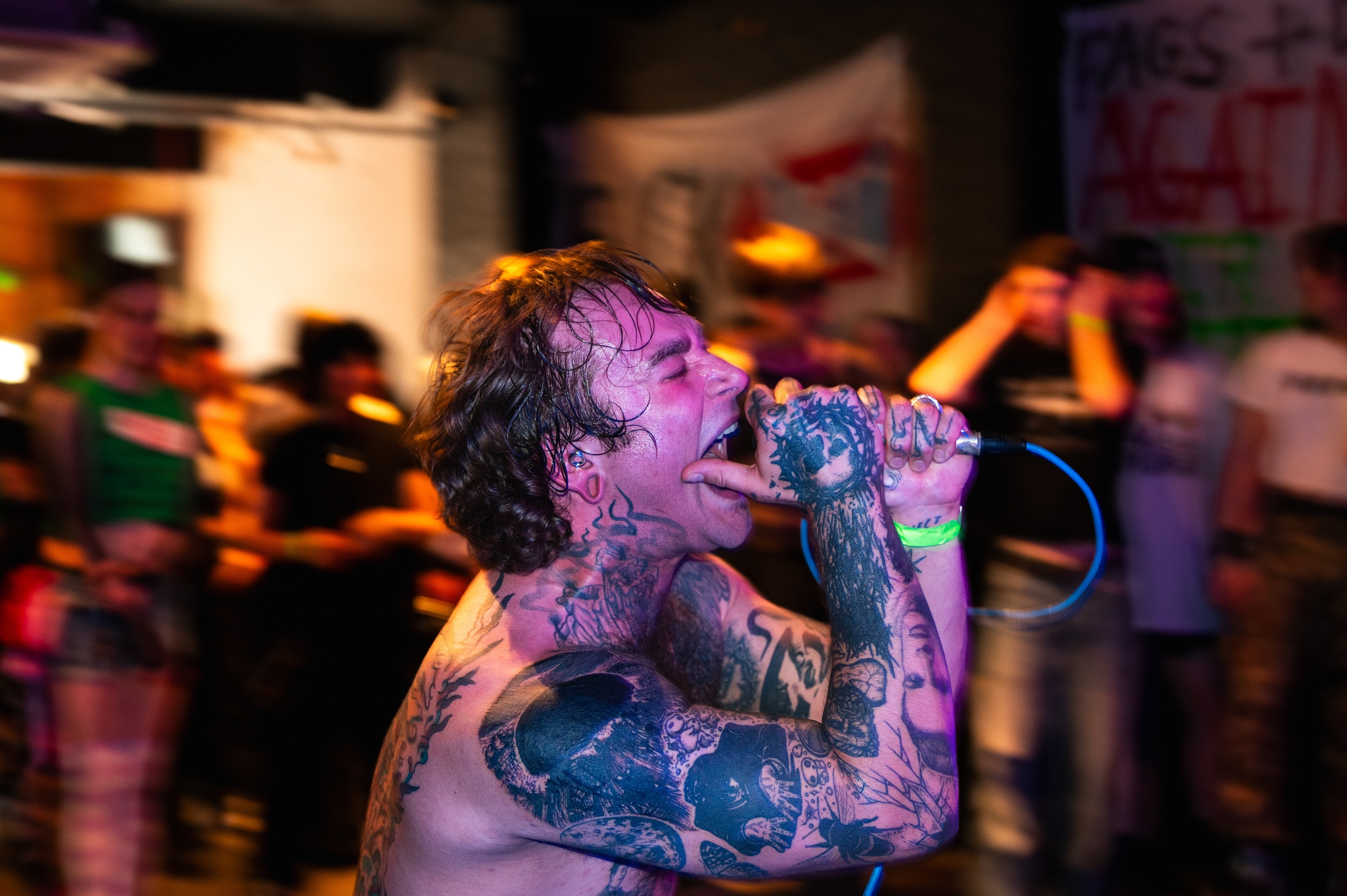 Tattooed performer singing into microphone under purple stage lighting, blurred crowd and venue in background.