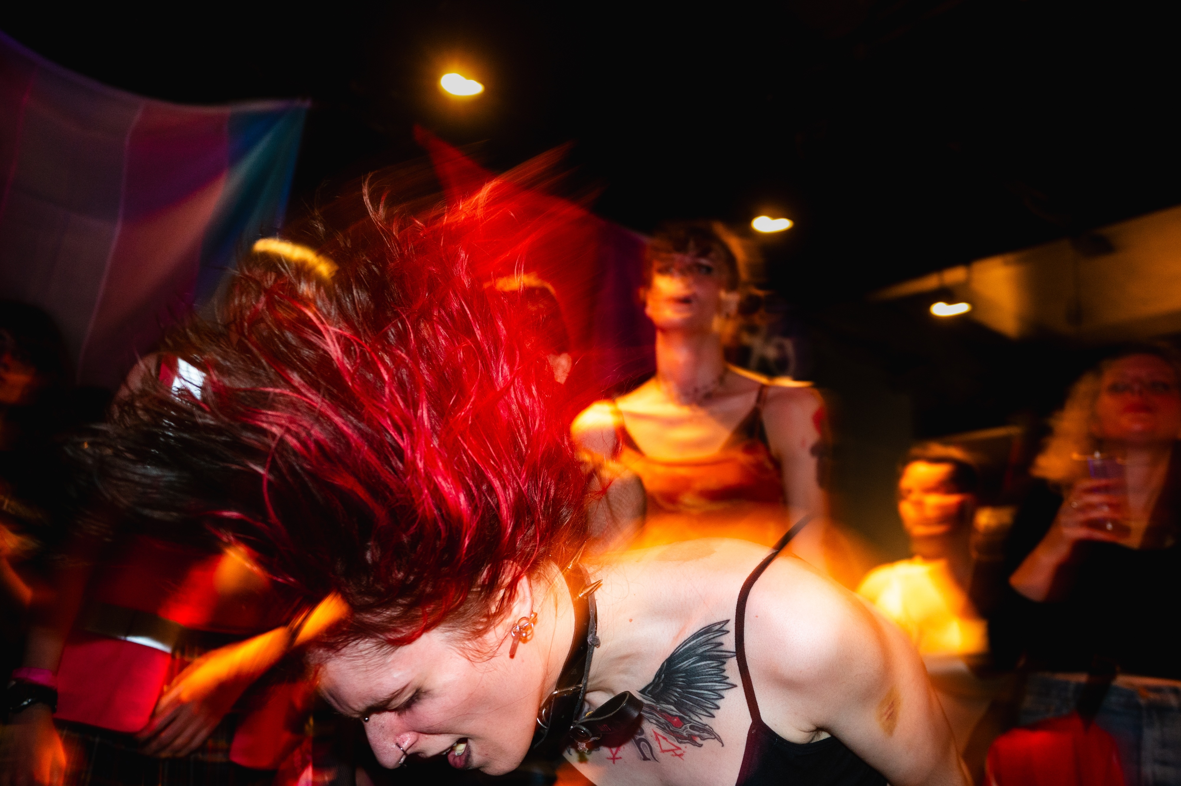 Person with spiky red hair headbanging in dimly lit venue with warm orange lighting and blurred crowd in background.