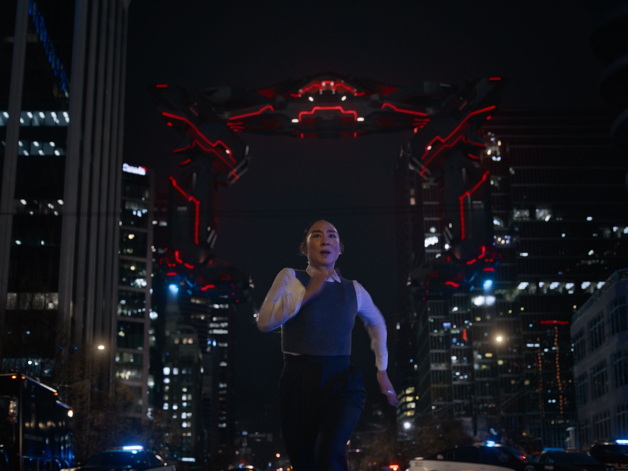Man in blue waistcoat and white shirt standing beneath large mechanical red tentacle-like appendages against illuminated city skyline at night.