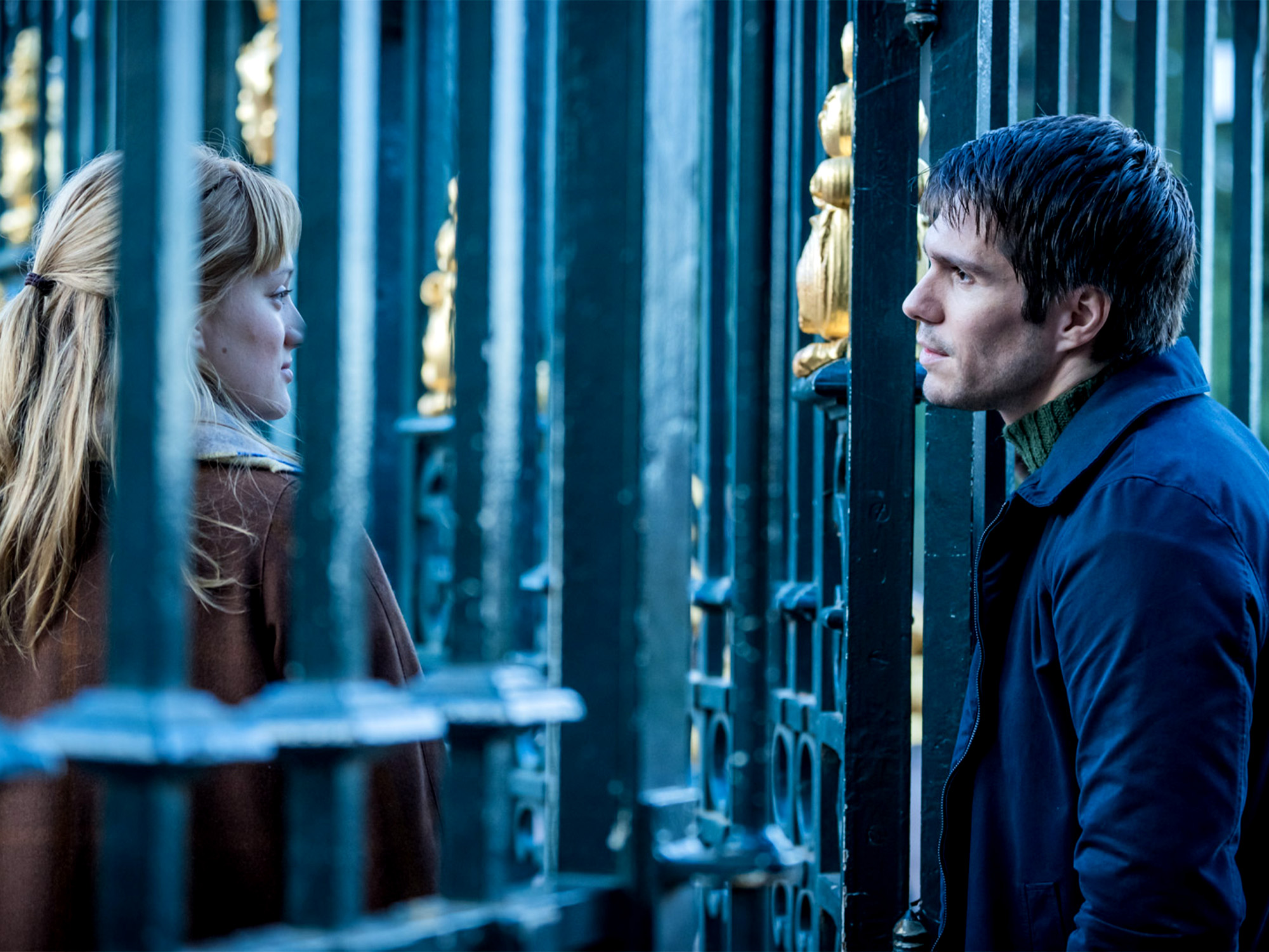 Blonde woman in brown jacket behind blue metal bars speaking to dark-haired man in blue coat standing outside the cell.