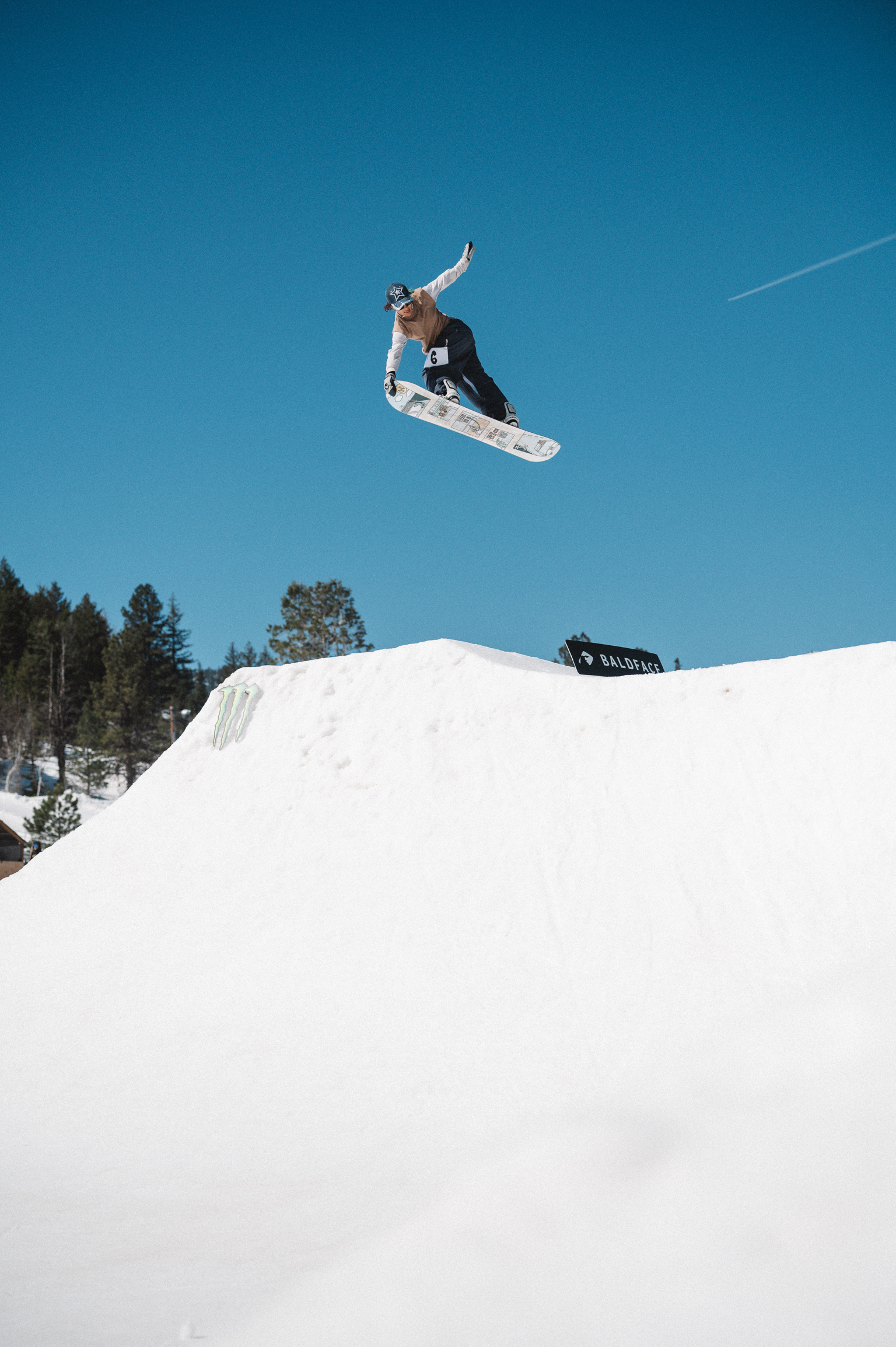 Snowboarder airborne above white snow ramp with evergreen trees and clear blue sky in background.
