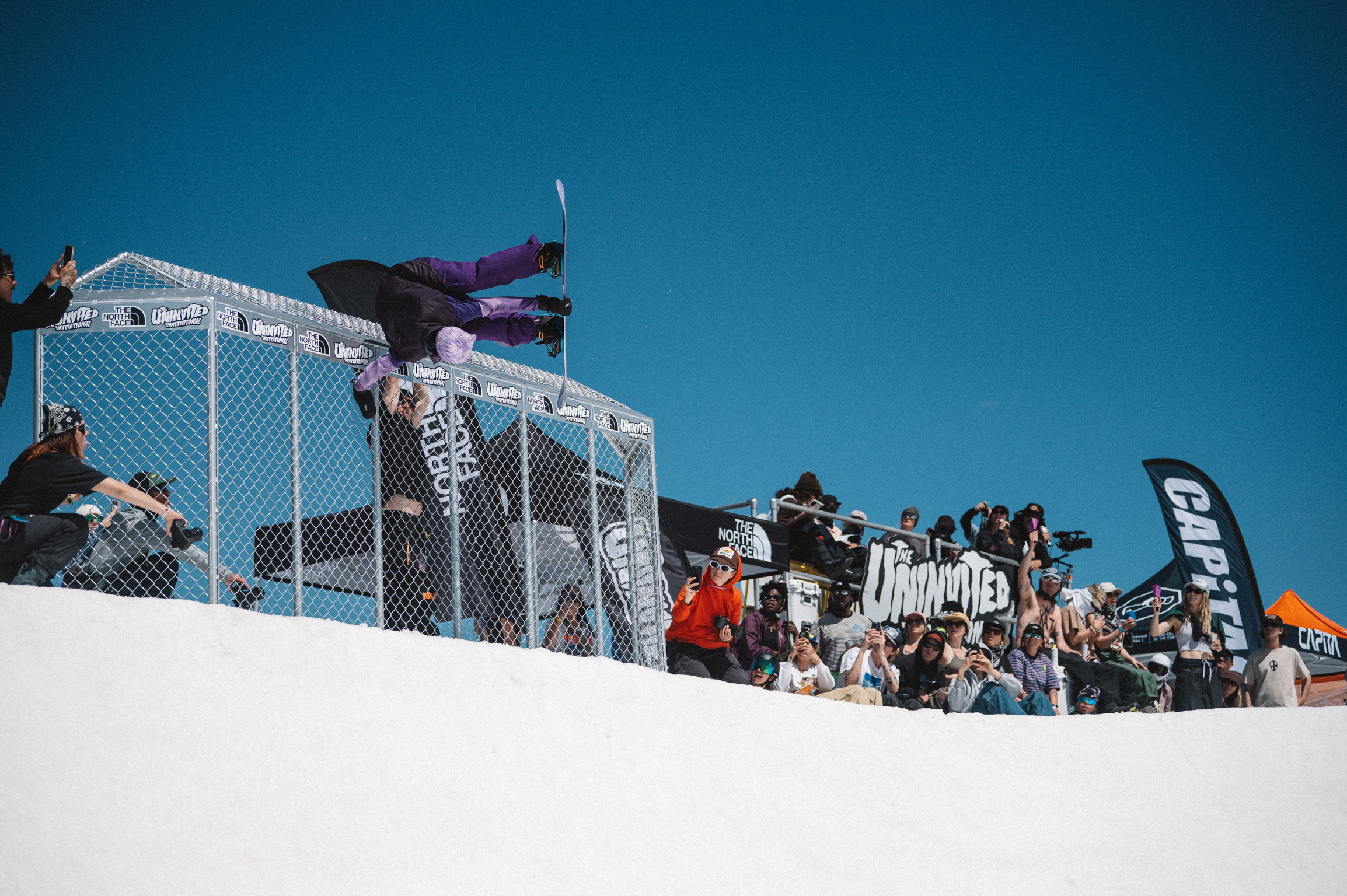 Spectators watching from metal viewing platform with chain-link fencing on snowy slope under clear blue sky.