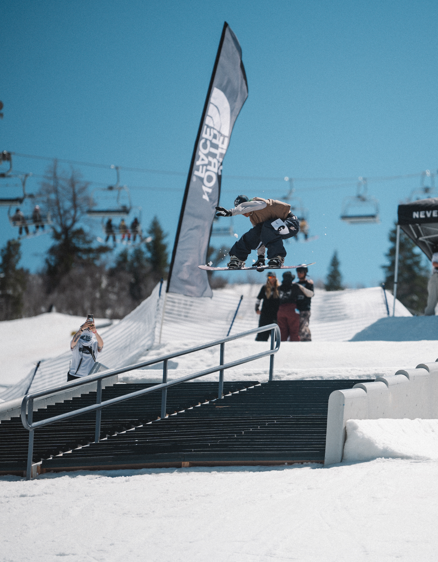 Snowboarder jumping over metal rail on snowy slope with grey banner, blue sky, trees and spectators in background.