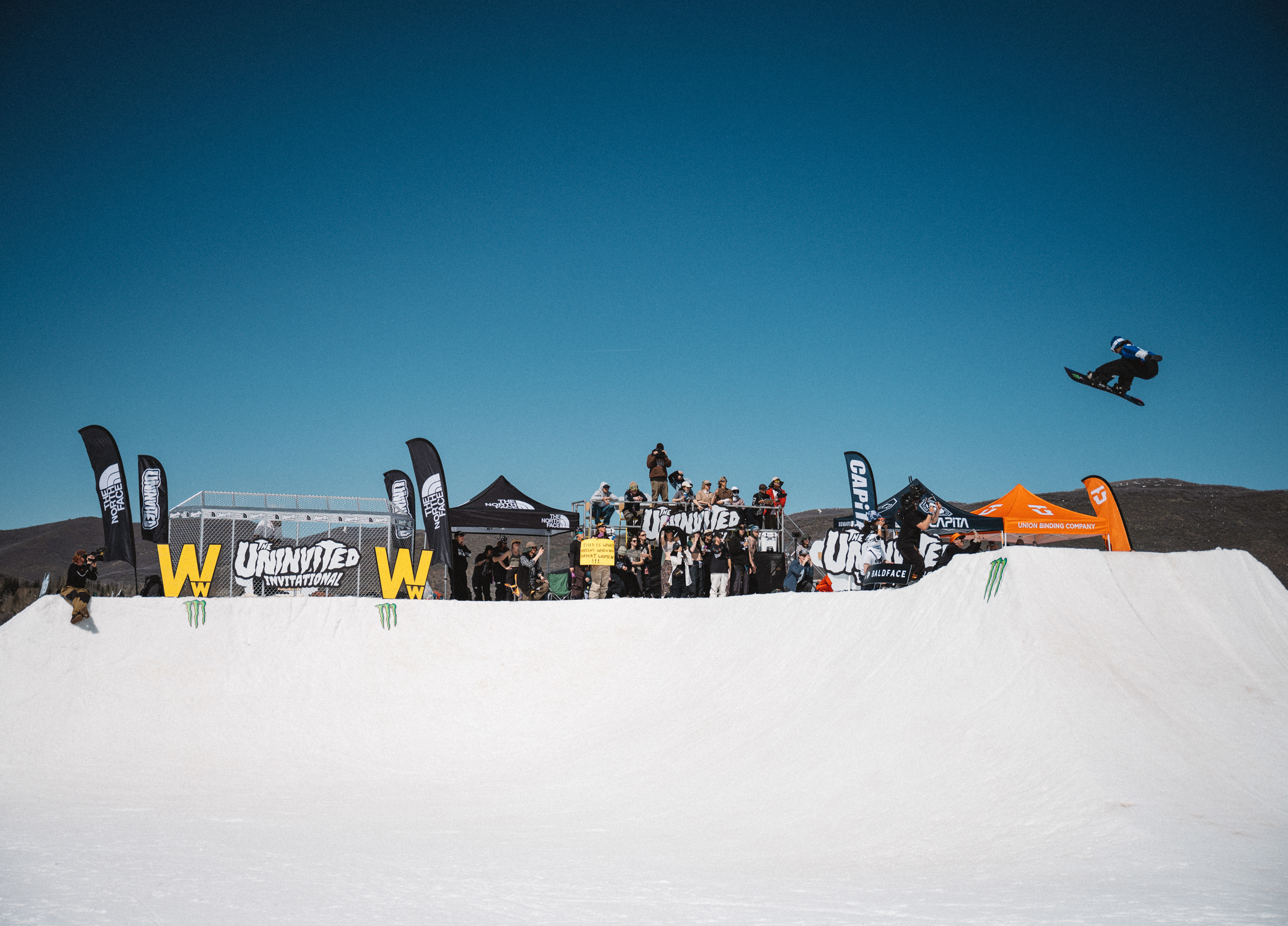 Snowboarder airborne above white halfpipe with spectators and sponsor banners including yellow "W" logos against clear blue sky.