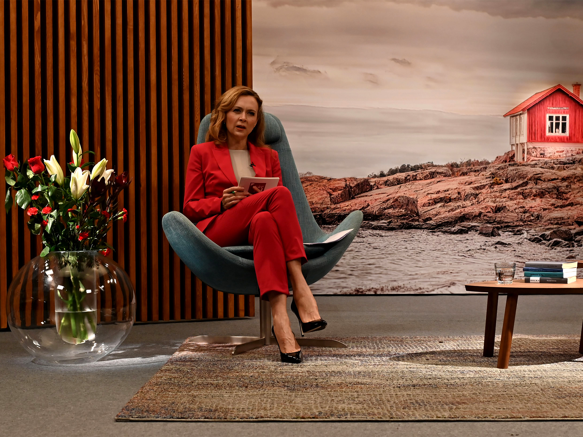 Woman in red suit sits in grey chair against wooden slats. Large coastal mural shows red house on rocky shore behind her.