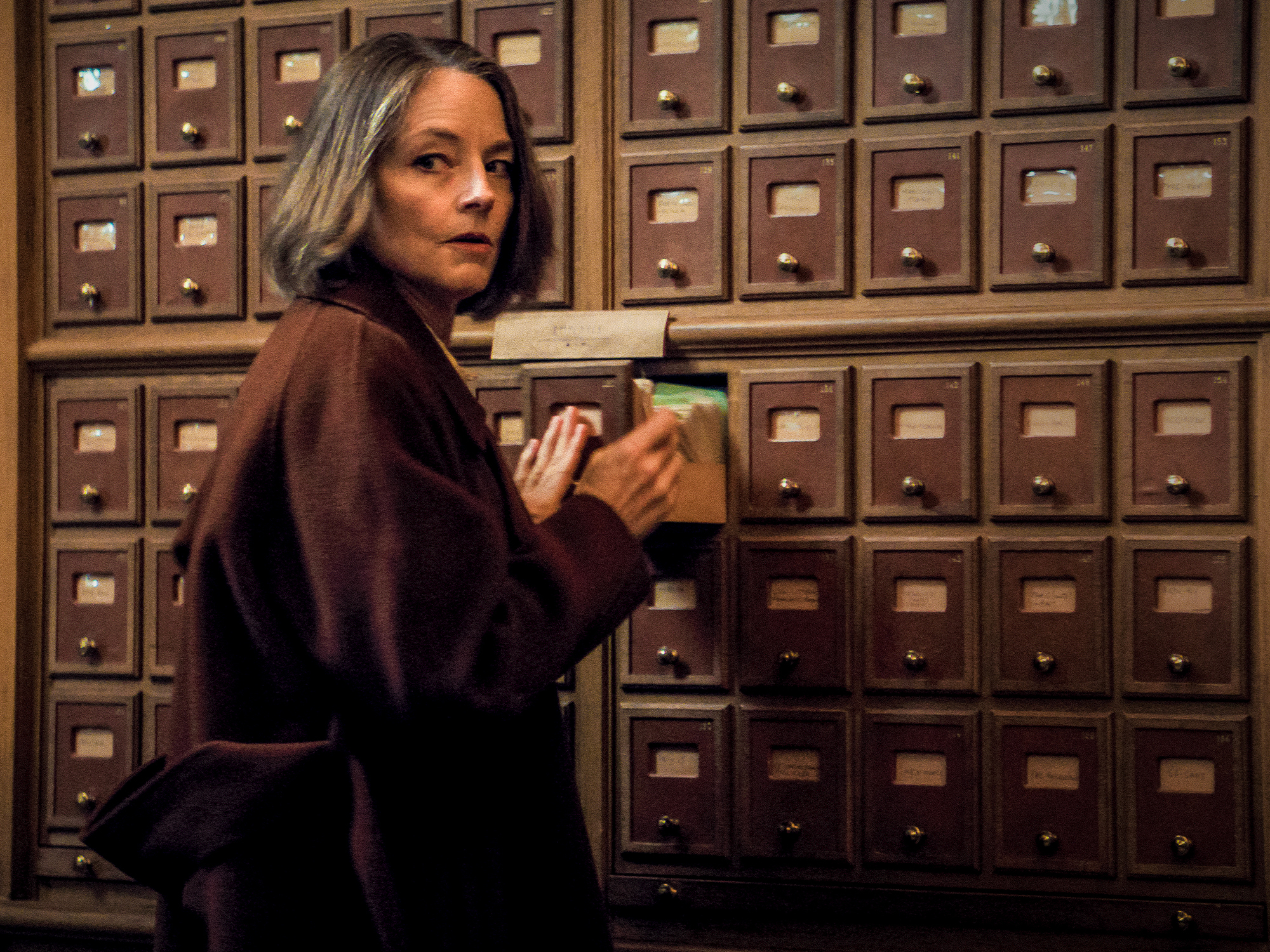 Woman in brown jacket standing in front of wall of cabinets.
