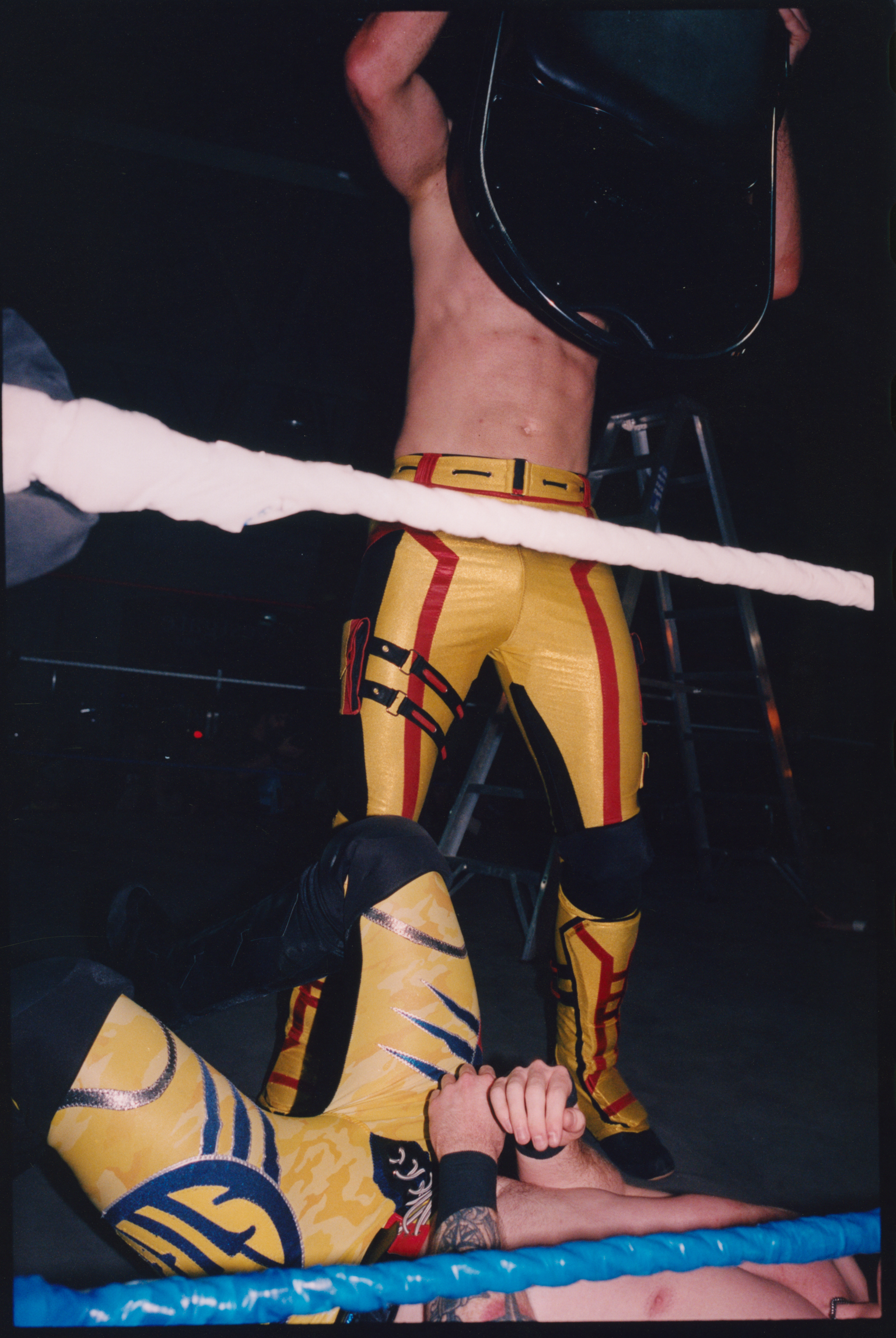 Two wrestlers in yellow and red gear grappling in ring, one standing over another lying on blue mat, white rope visible.