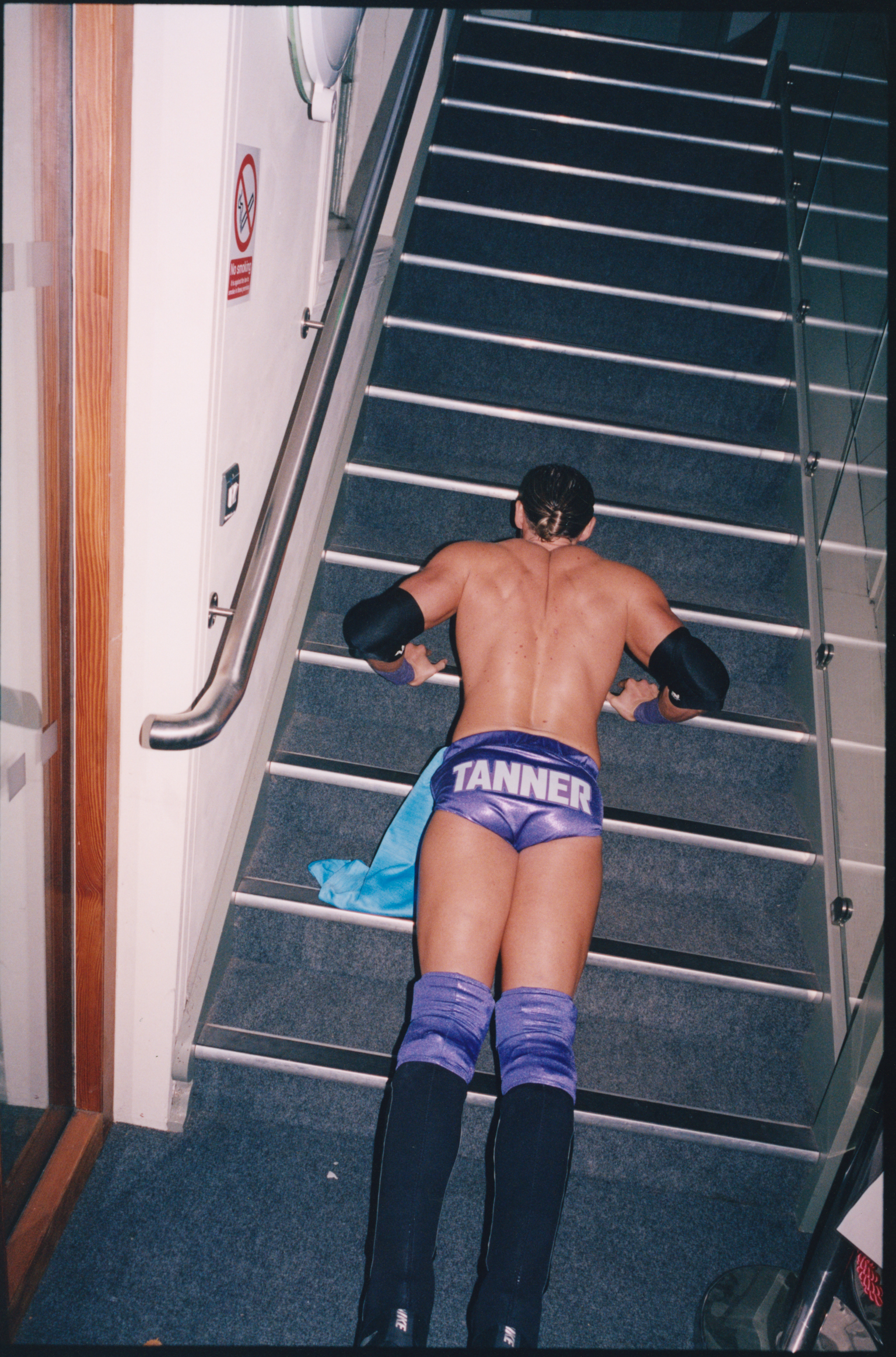 Person in purple wrestling trunks with "TANNER" text and purple socks doing push-ups on ship stairs with metal railings.