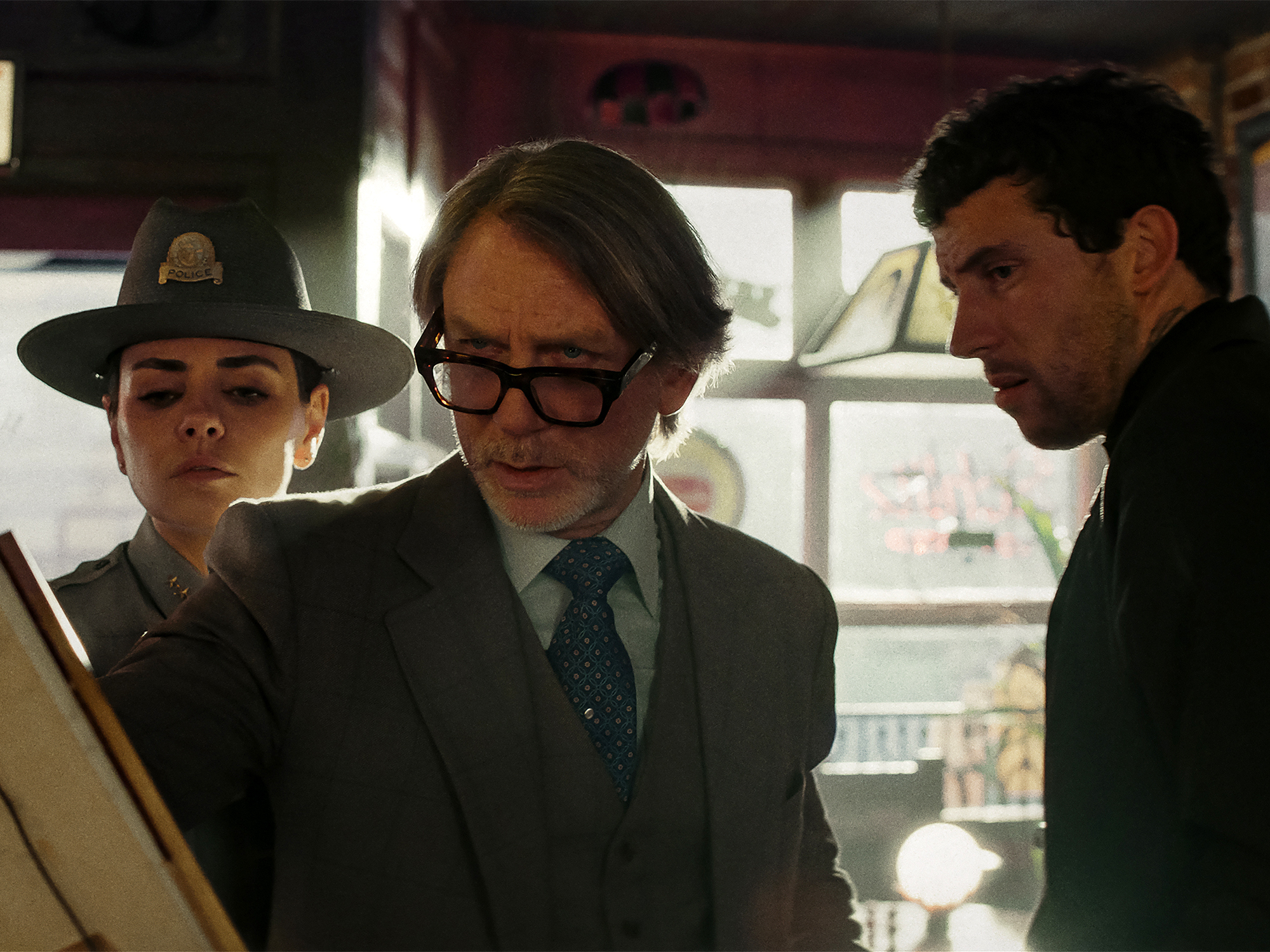 Three people in dark clothing stand inside what appears to be a vehicle or confined space. One wears a hat, another has glasses and a tie.