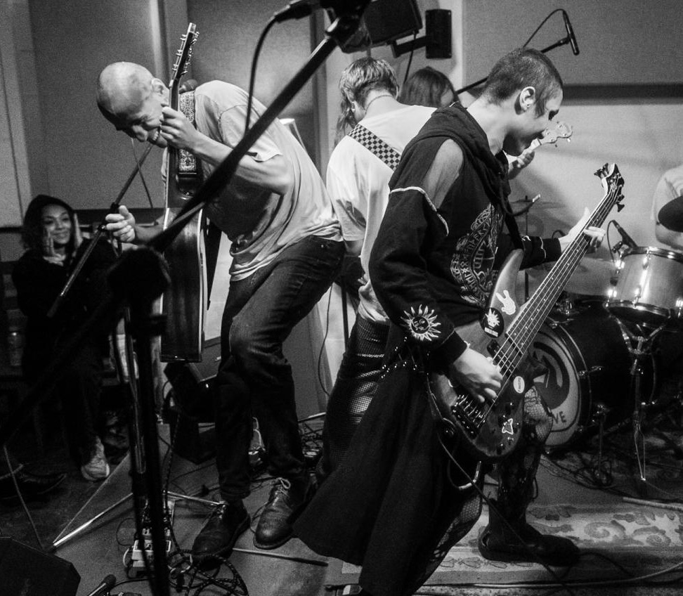 Black and white image of three musicians performing on stage - vocalist at microphone, bassist, and drummer, with audience members visible in background.