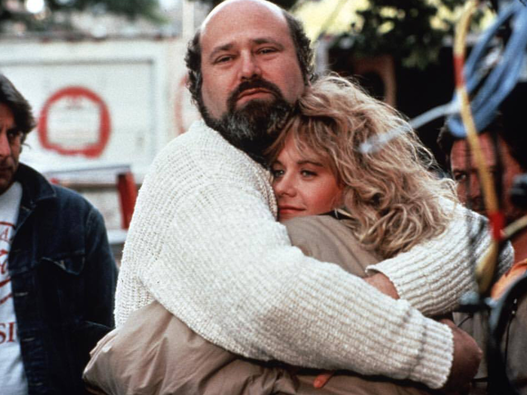 Bald man with beard embracing blonde woman in white jumper on busy street with blurred background of people and signs.