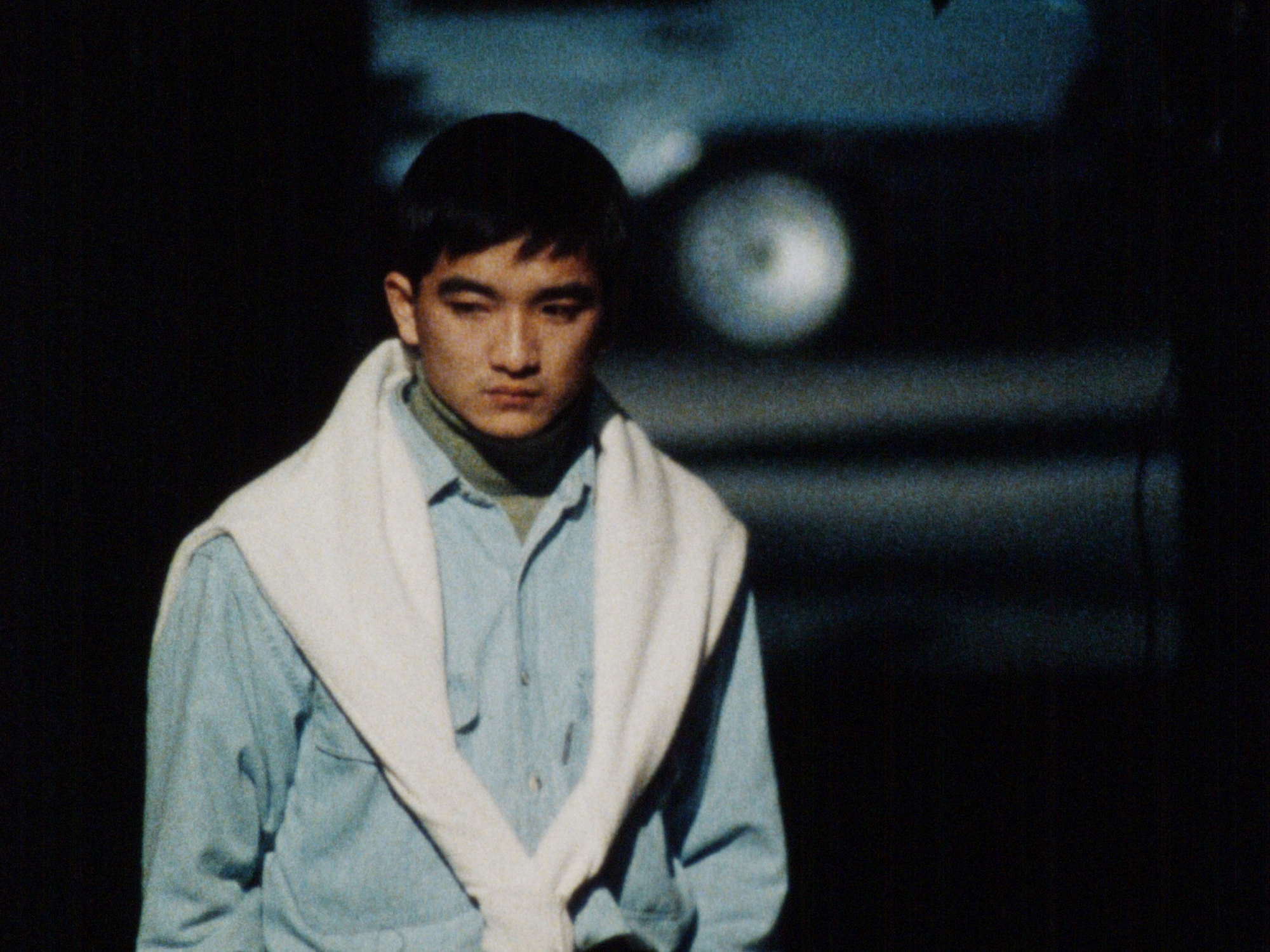 A young East Asian man wearing a blue shire with a white sweater around his shoulders stands on a street with an uncertain expression.