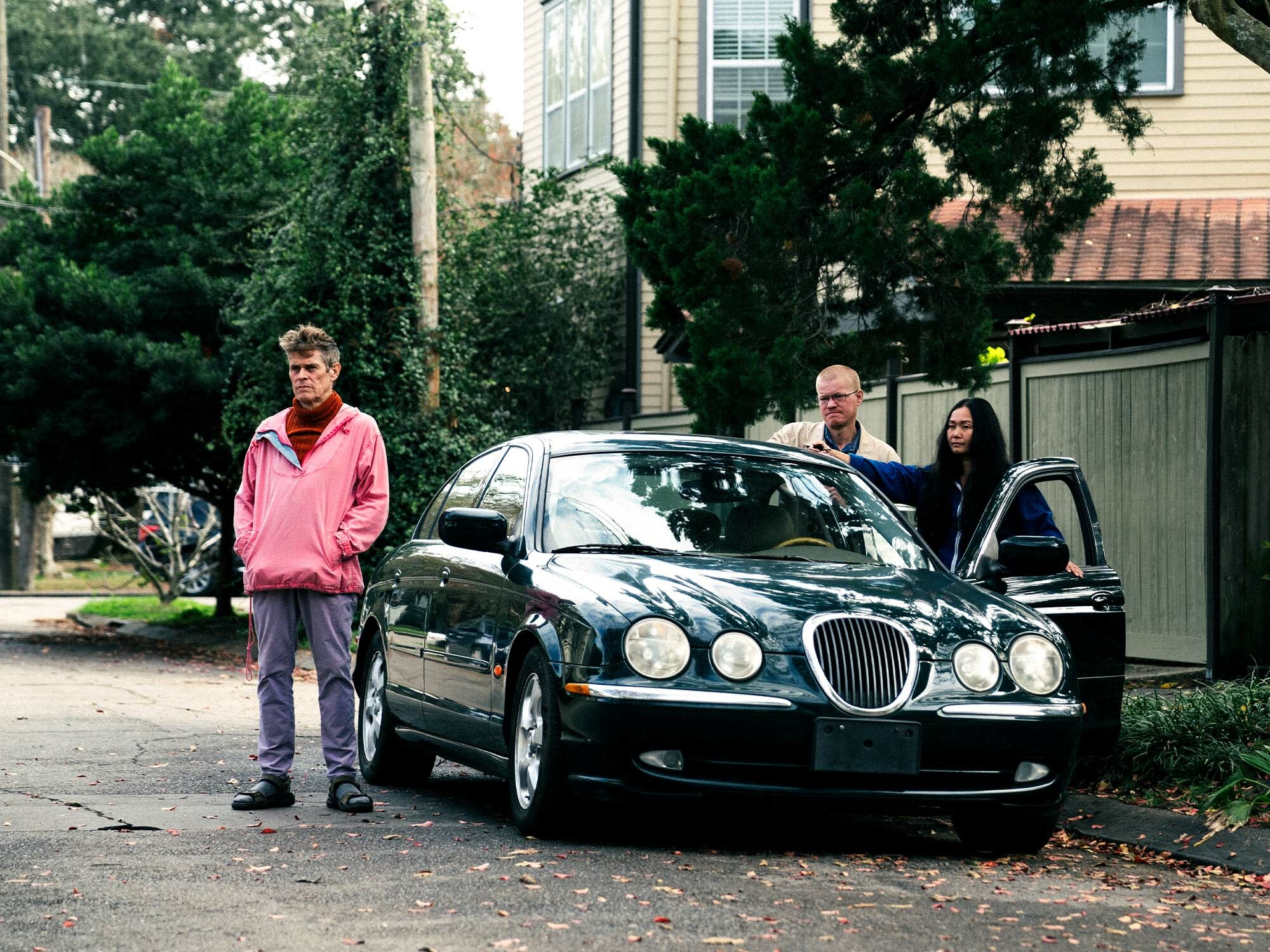 Suburban street with trees and houses; parked black car; three people standing near the car, two men and one woman.
