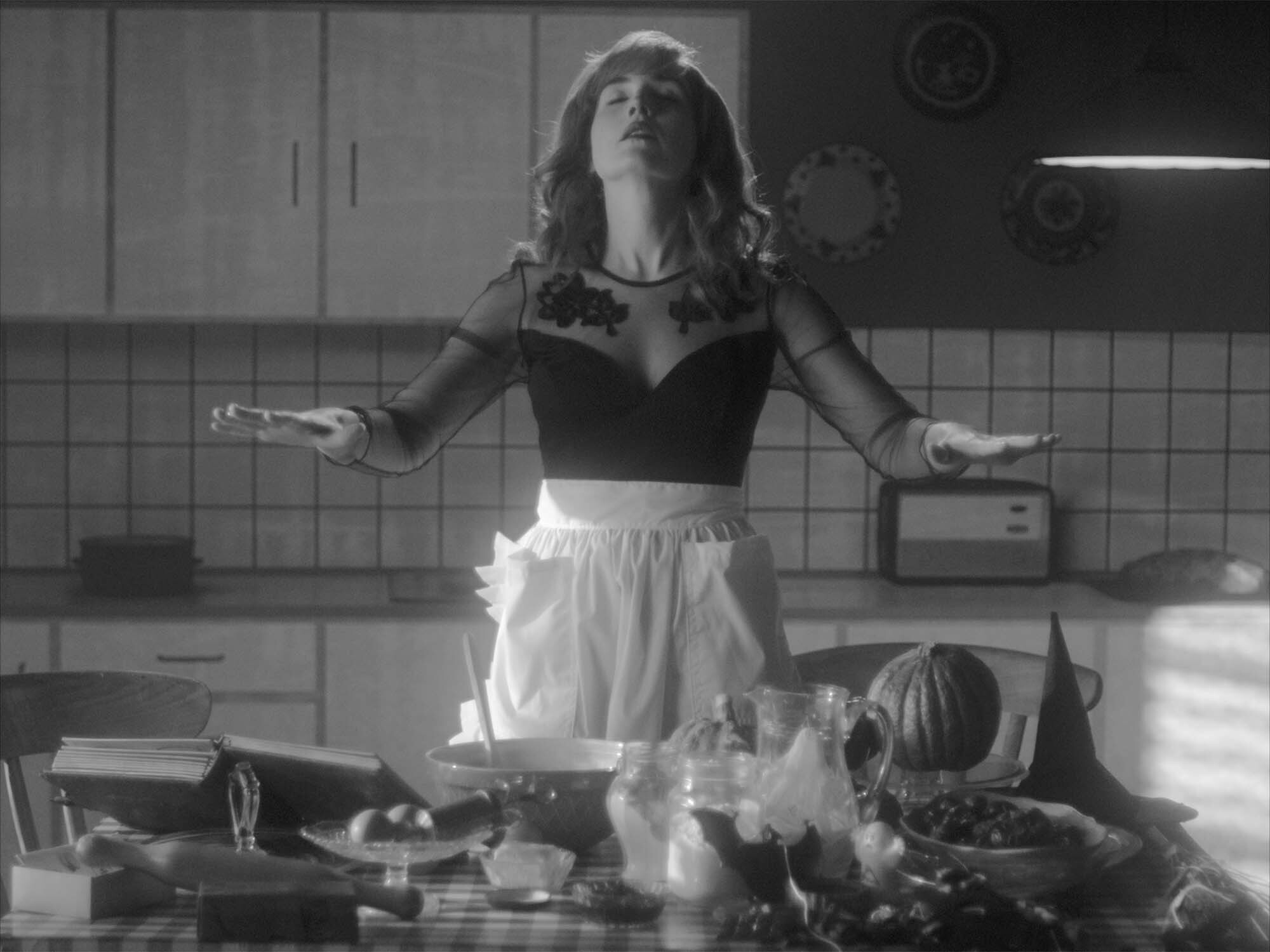 Woman in black top and white skirt stands amidst cluttered kitchen