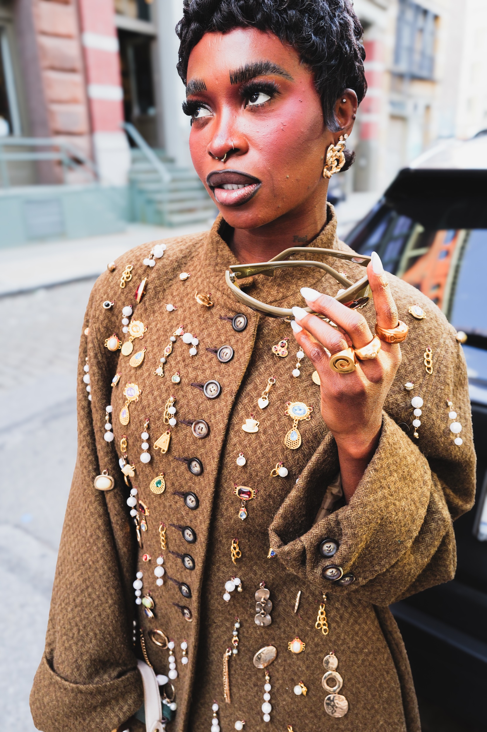 Woman with short dark hair wearing brown coat decorated with gold buttons, pins and embellishments, holding sunglasses on city street.