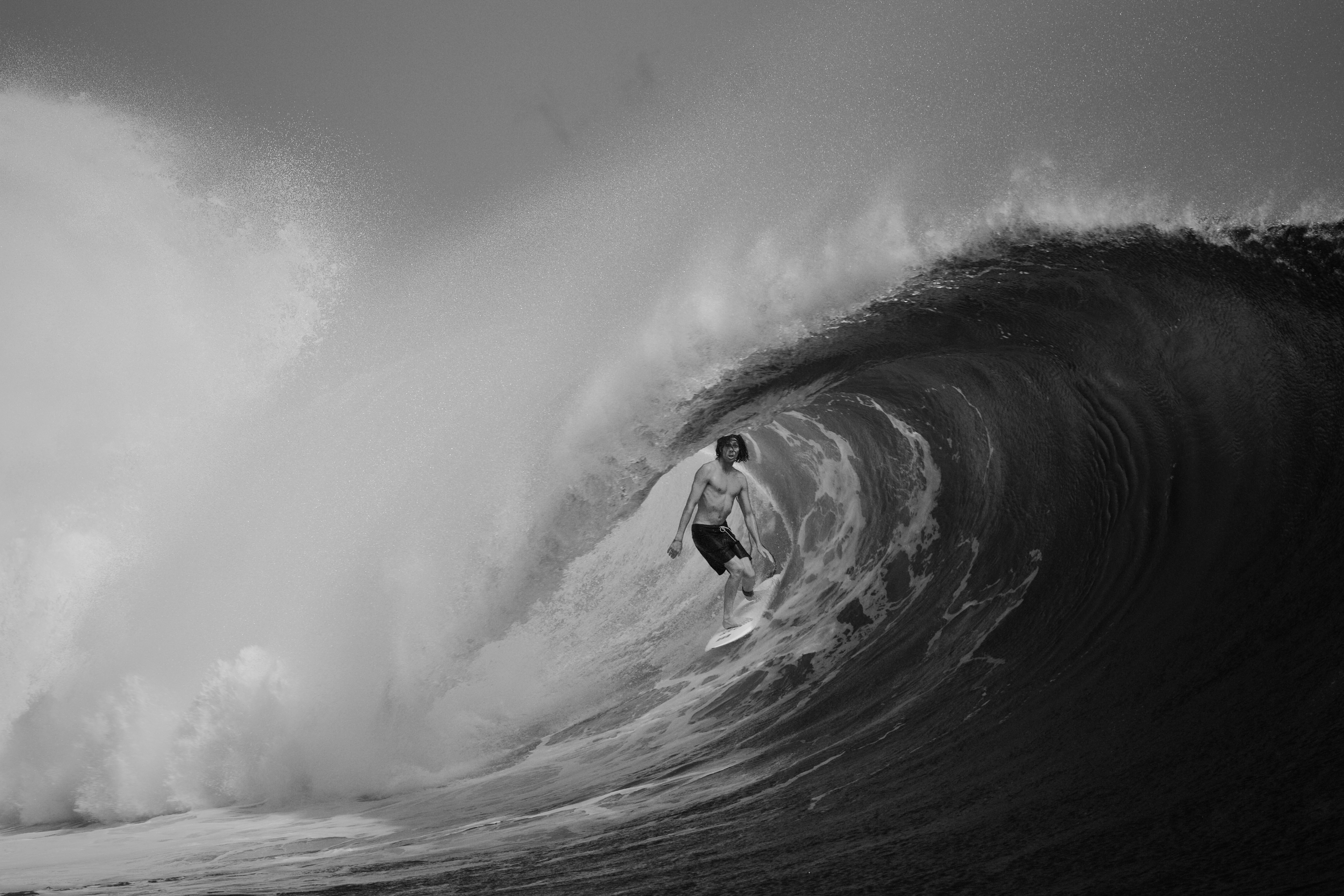 Large ocean wave curling to form barrel shape with surfer visible inside, captured in black and white with dramatic spray and foam.