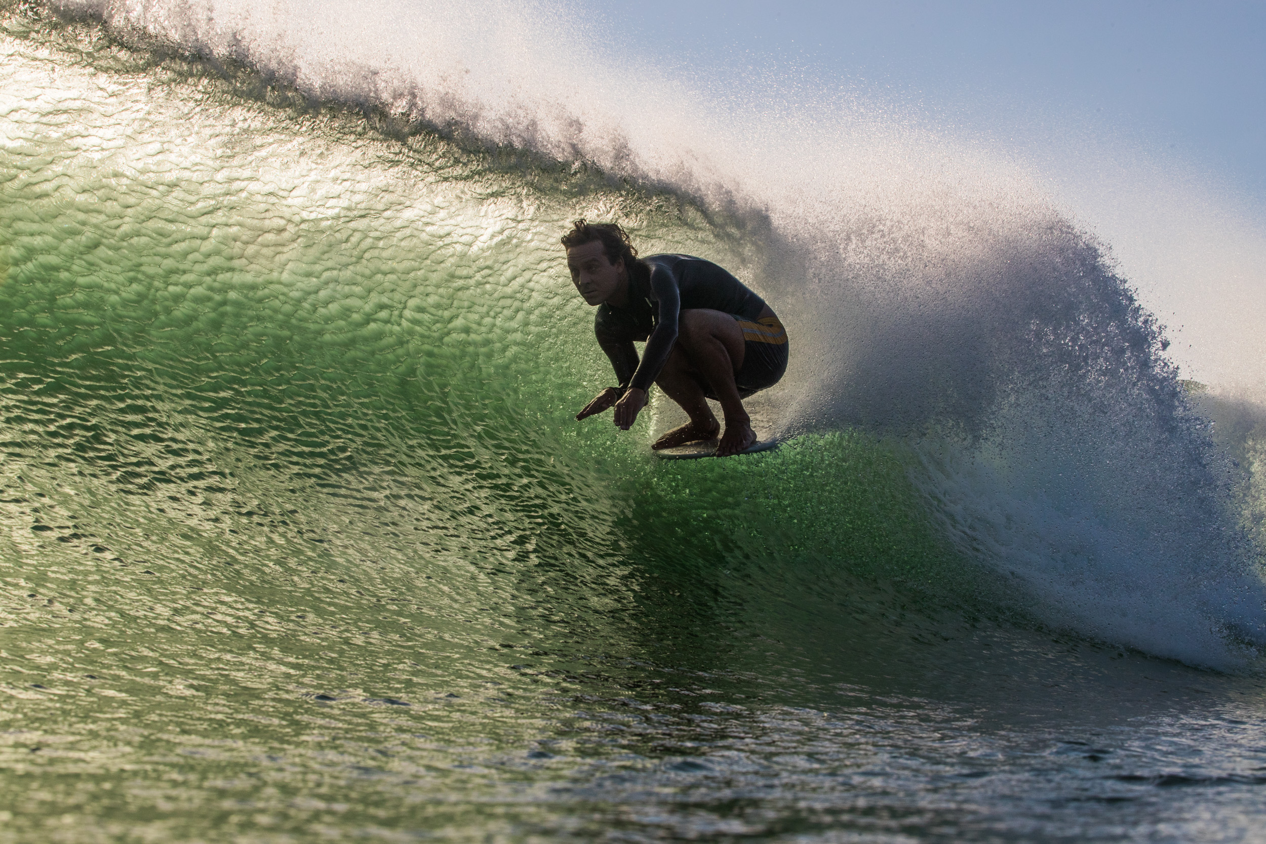 Surfer crouched on surfboard inside green wave barrel with white foam spray and blue sky visible above