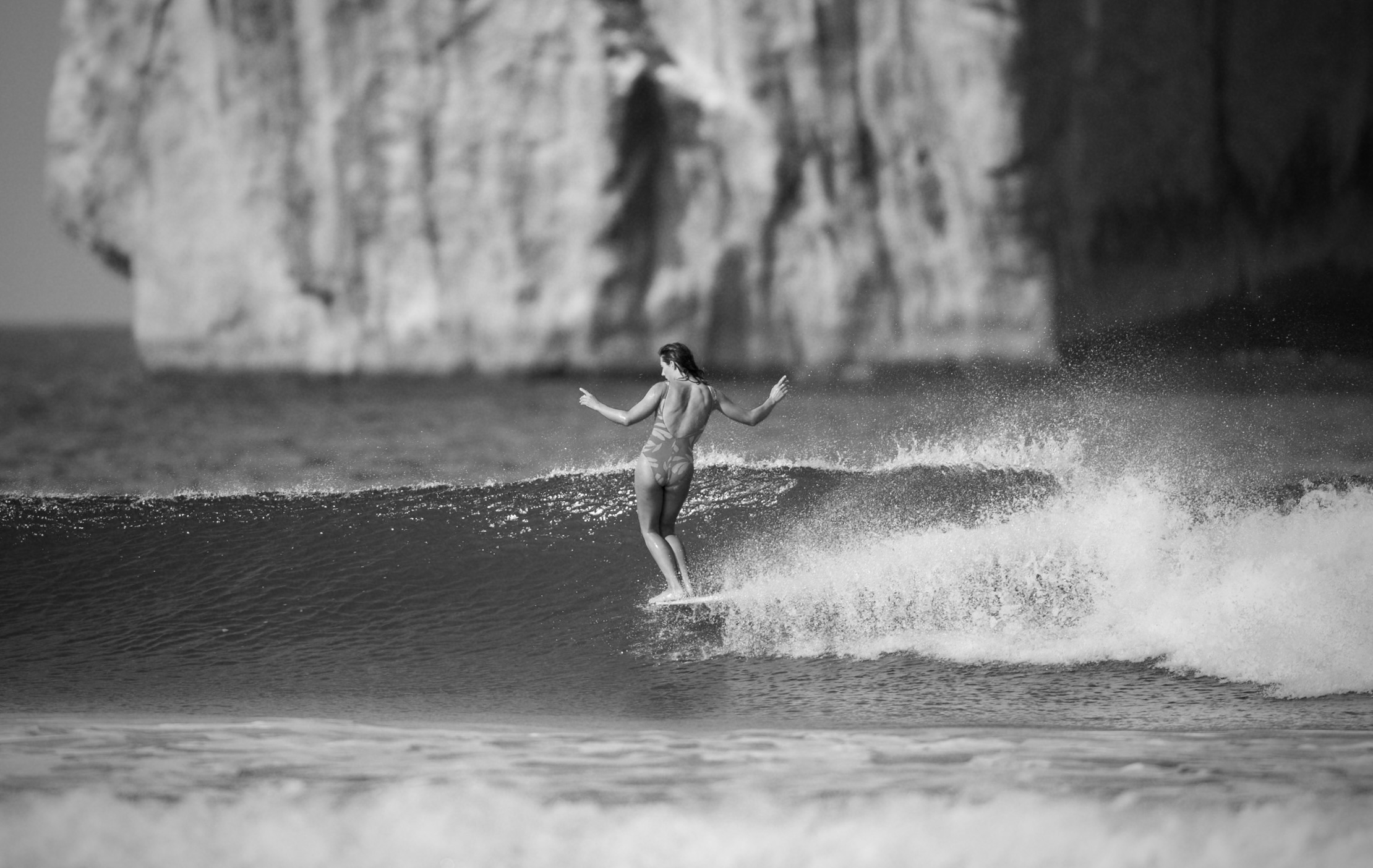 Black and white image of surfer riding wave with arms outstretched, rocky cliff face in background, white foam spray around surfboard.
