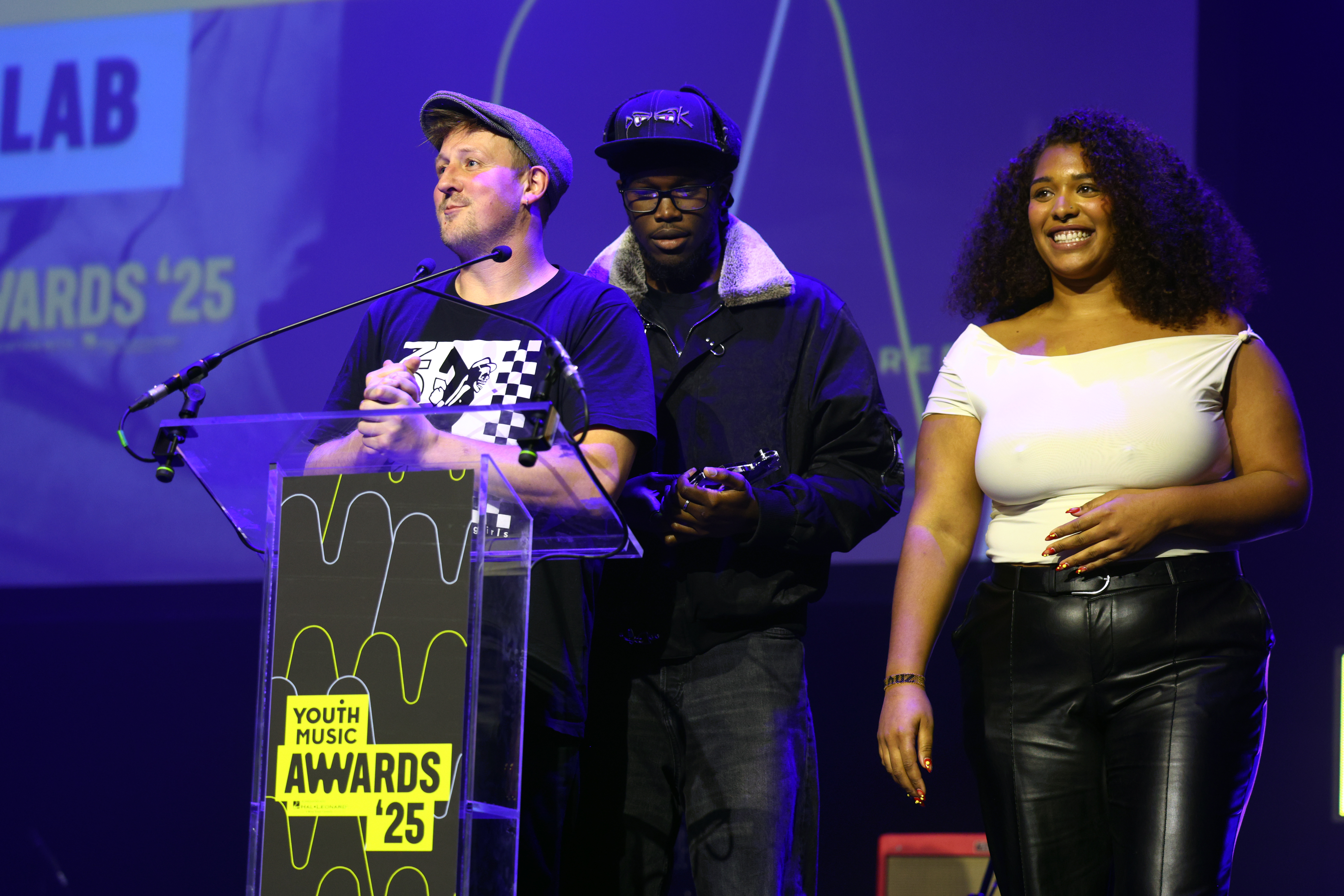 Three people on stage at Youth Music Awards podium. Man in cap at microphone holding award, flanked by man in hat and woman in white top.