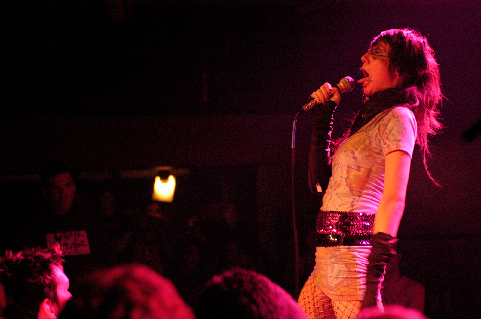 Female performer singing into microphone on stage with pink lighting, wearing white top and patterned skirt, crowd visible below.