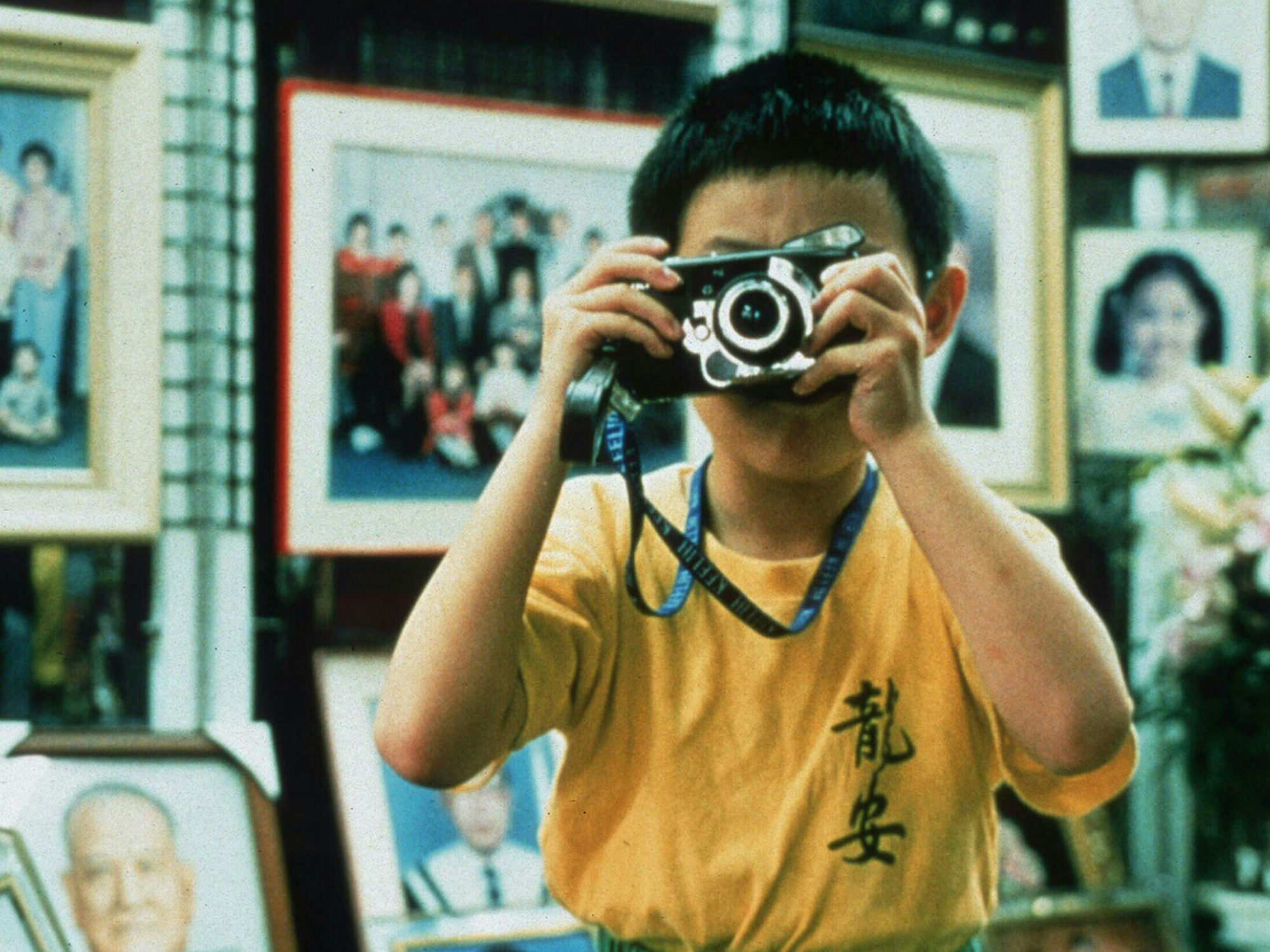 A young Taiwanese boy in a yellow shirt holding a camera in a room with framed pictures on the walls.
