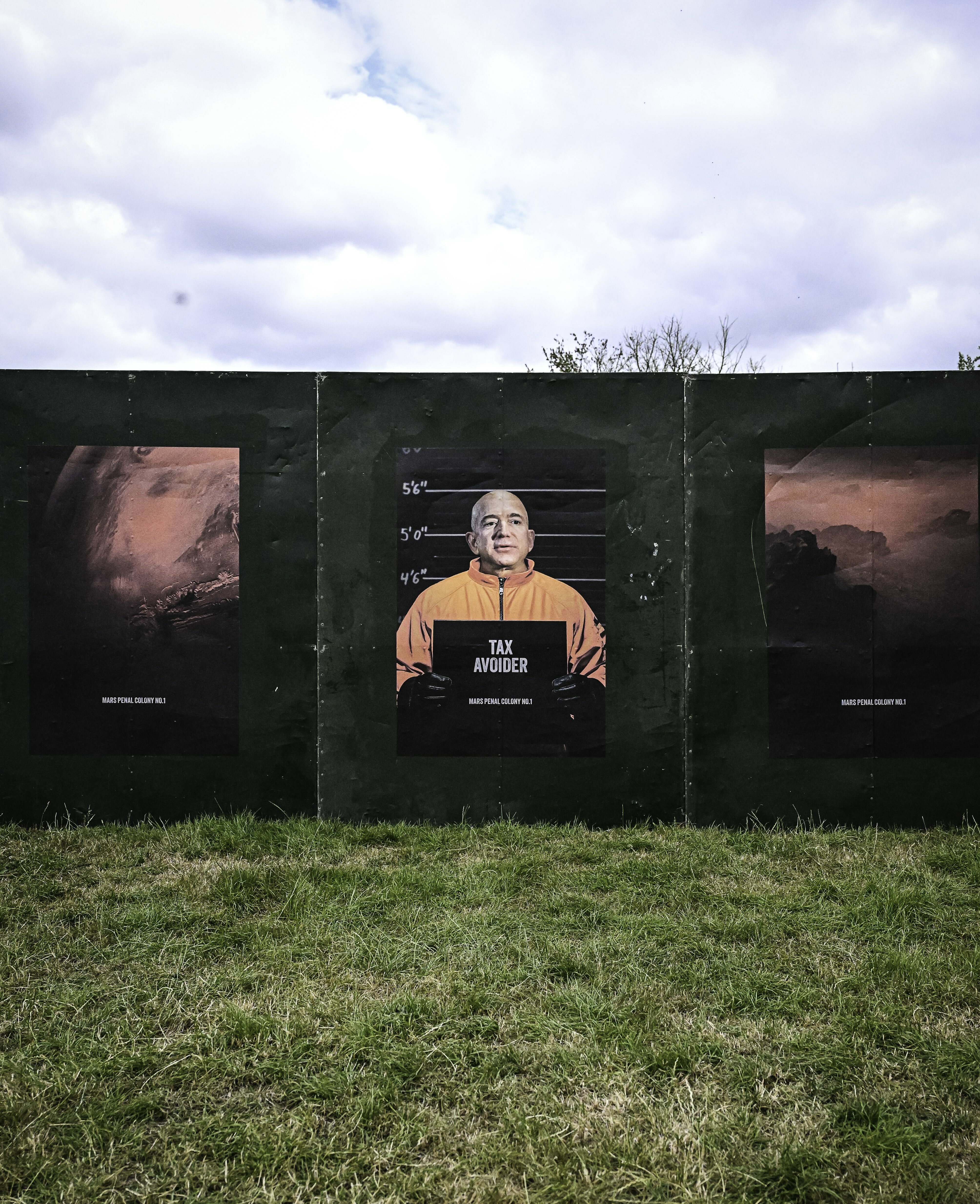Man in orange prison uniform behind bars on green wall mural, flanked by pink-toned landscape paintings on grass field.
