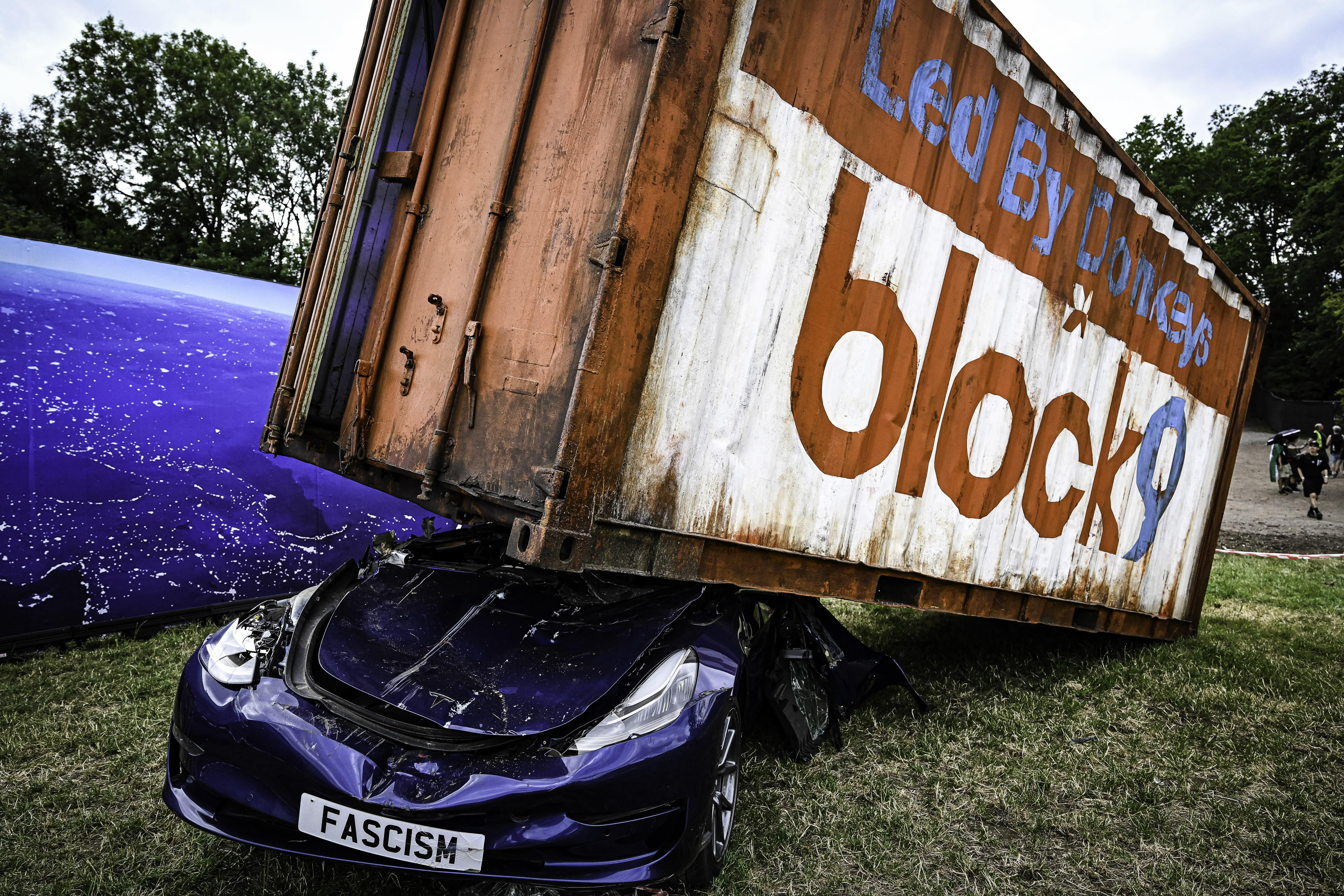 Blue car with "FASCISM" numberplate crushed beneath large orange and white Blockbuster video shop sign outdoors on grass.