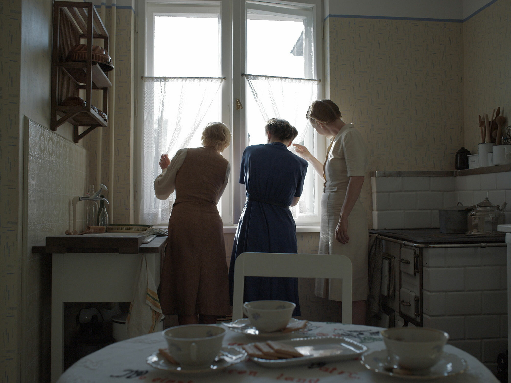 Three women in 1940s clothing stand by a window in a domestic interior.