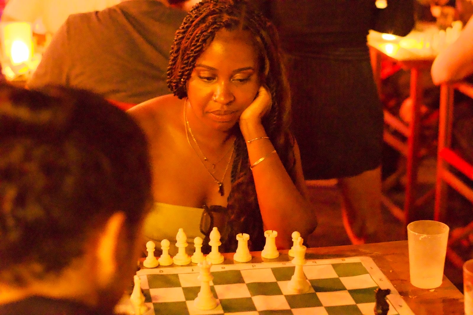 Woman with braided hair resting chin on hand, contemplating chess board with white pieces. Warm orange lighting, glass nearby.