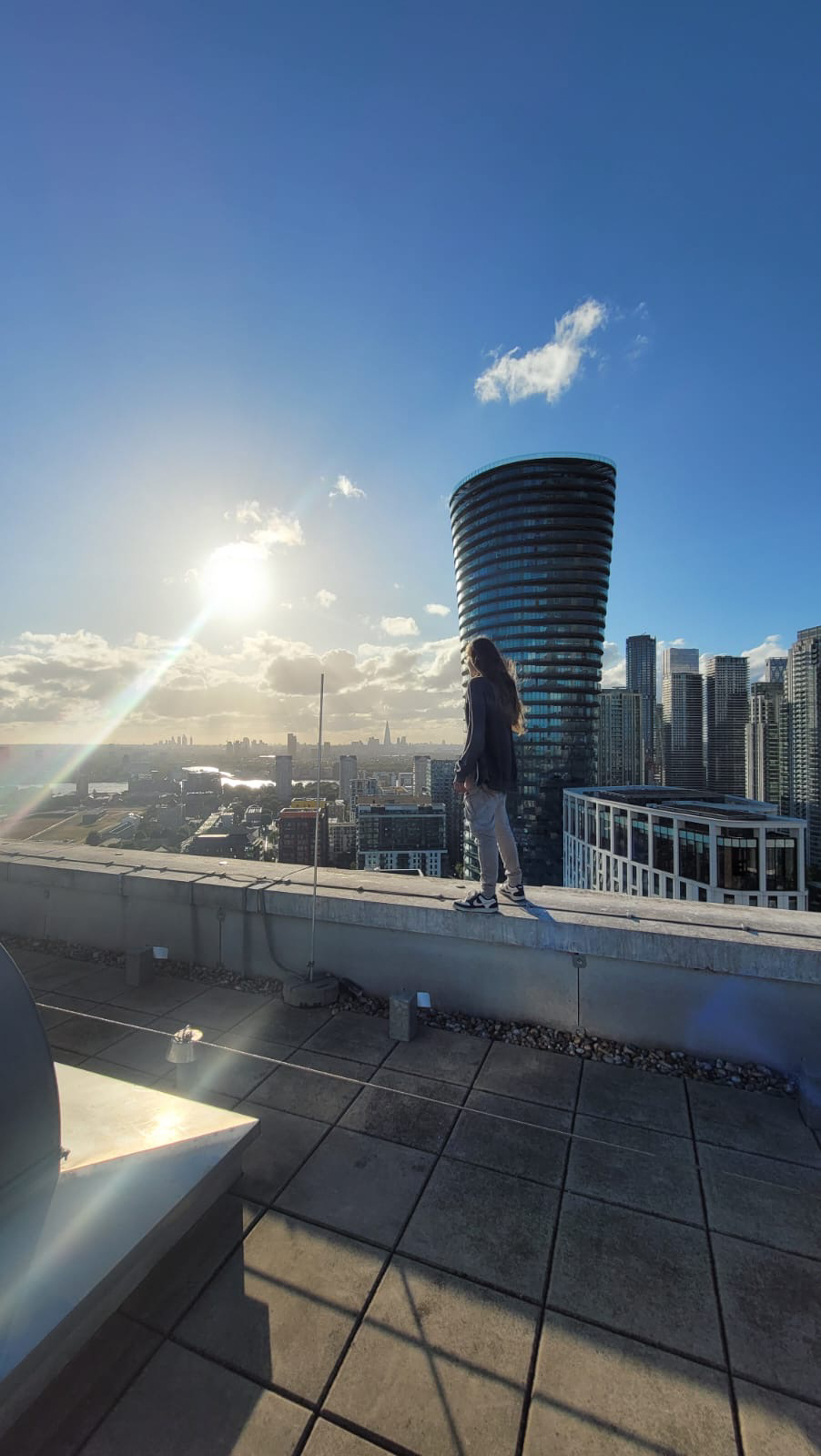 Person standing on rooftop terrace overlooking city skyline with tall buildings, bright sun creating lens flare against blue sky.